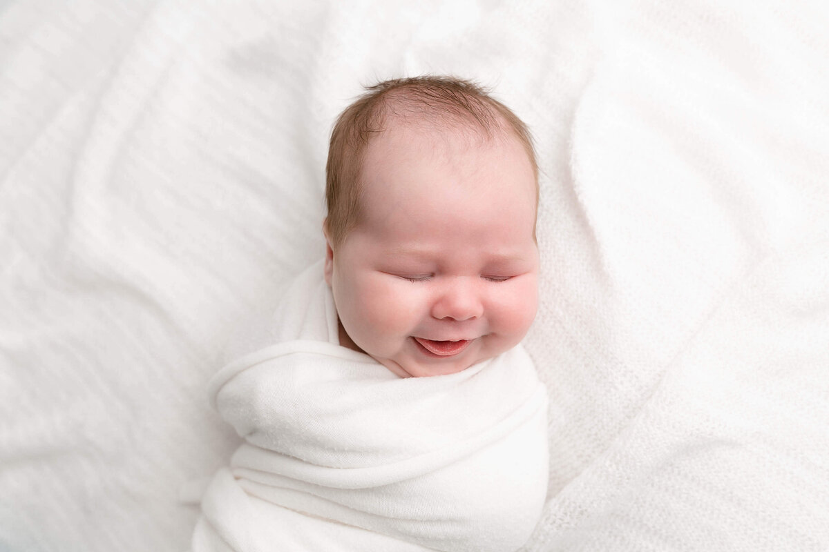 Newborn baby swaddled in white and laying on a white textured blanket. Baby has eyes closed and a big smile on her face and her tongue is sticking out a little bit.