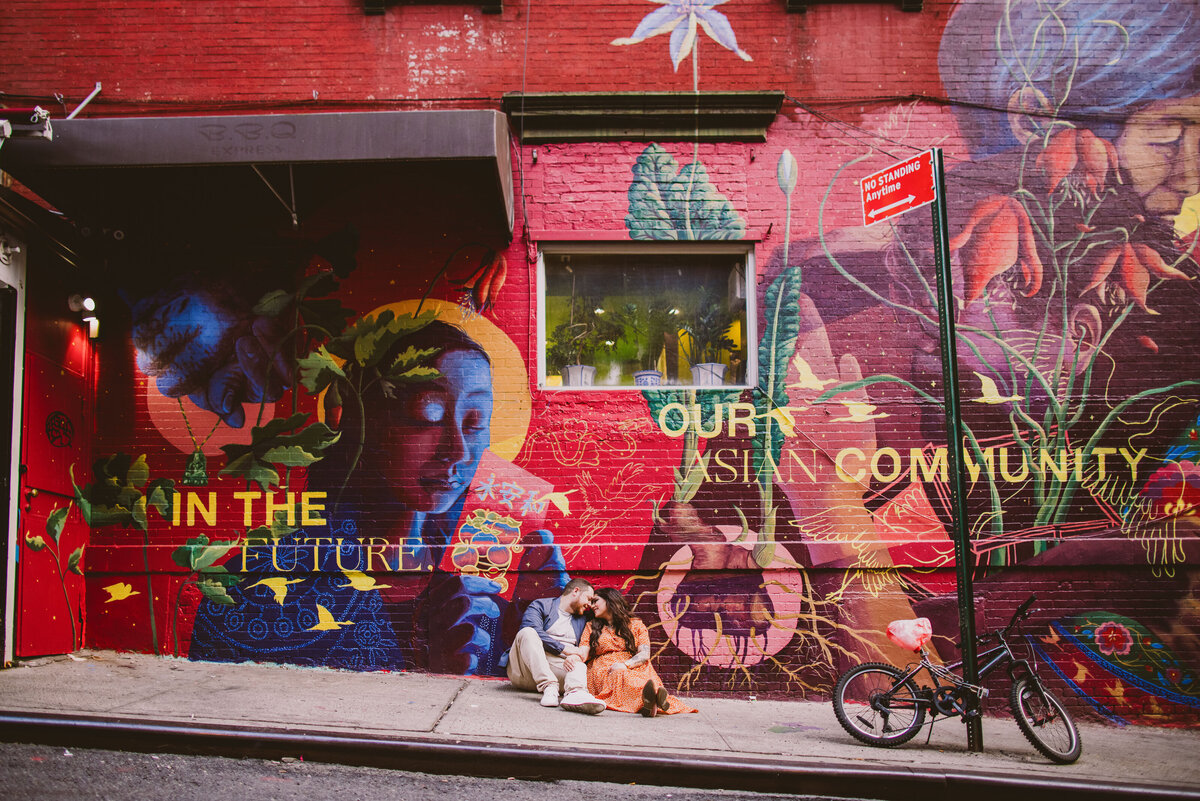 A couple sitting on a street in front of wall art in chinatown nyc during their engagement session