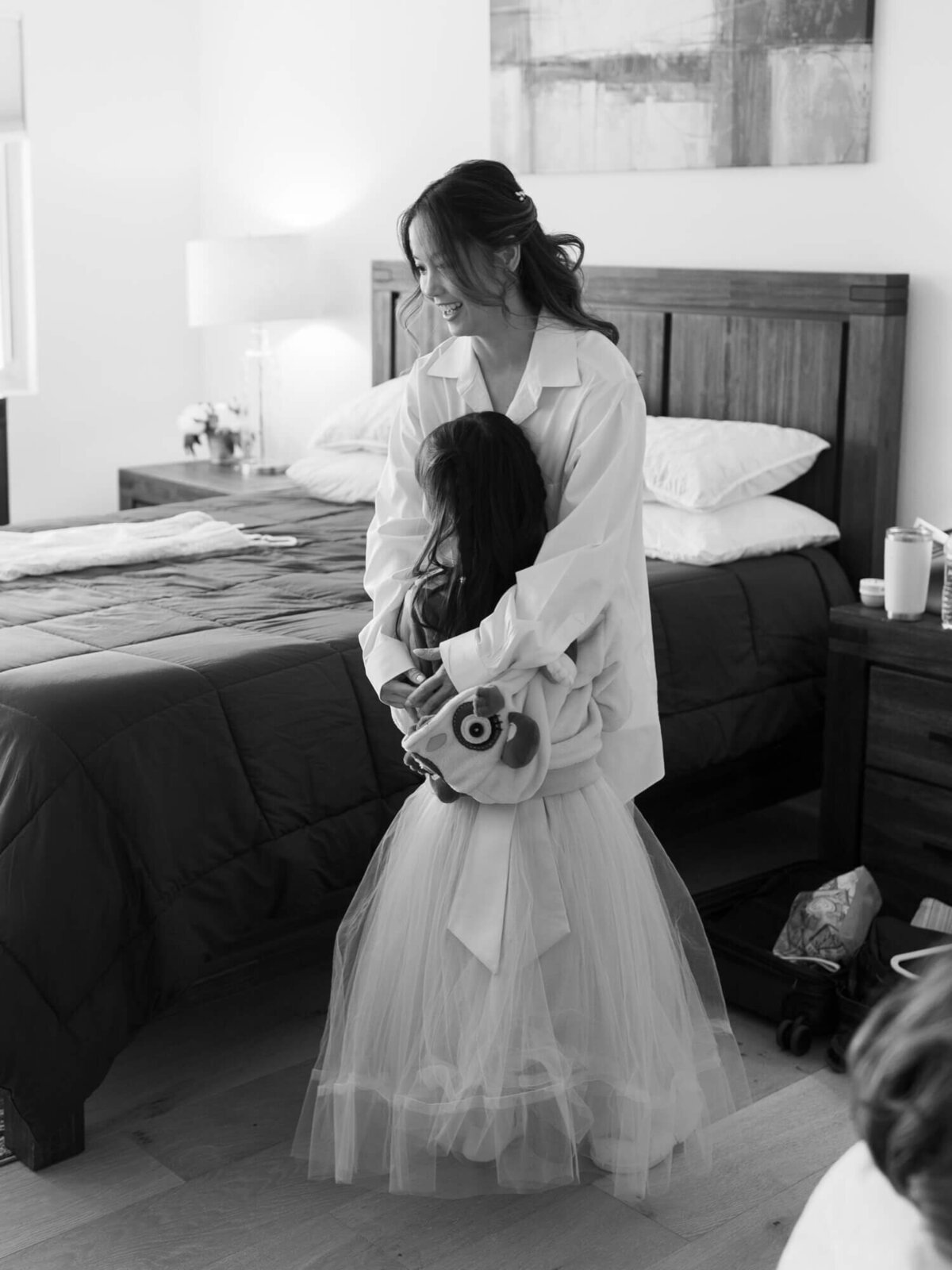 Bride and flower girl share a joyful hug in a bedroom.
