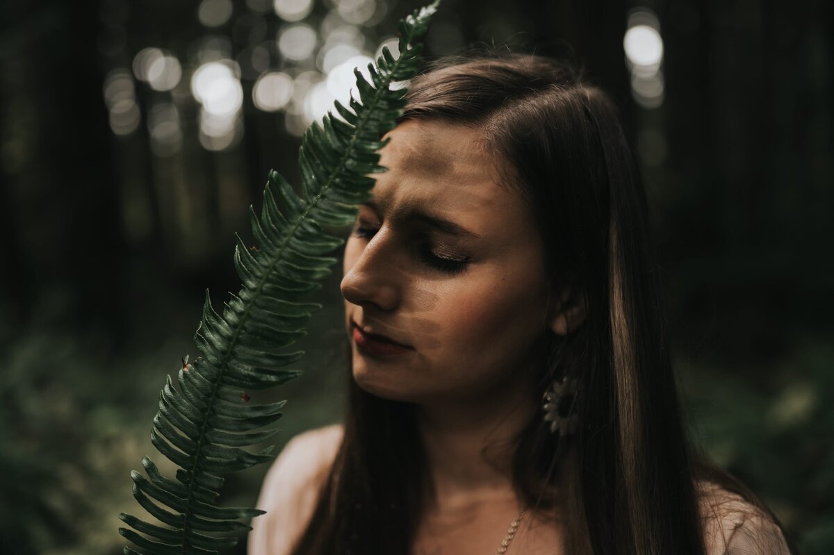 Woman closes her eyes and holds fern in front of her face in the forest.