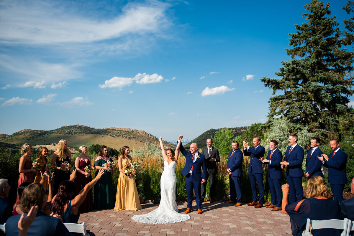 A wide angle shot of a bride and groom and their wedding party as the bride and groom throw their hands in the air in celebration.