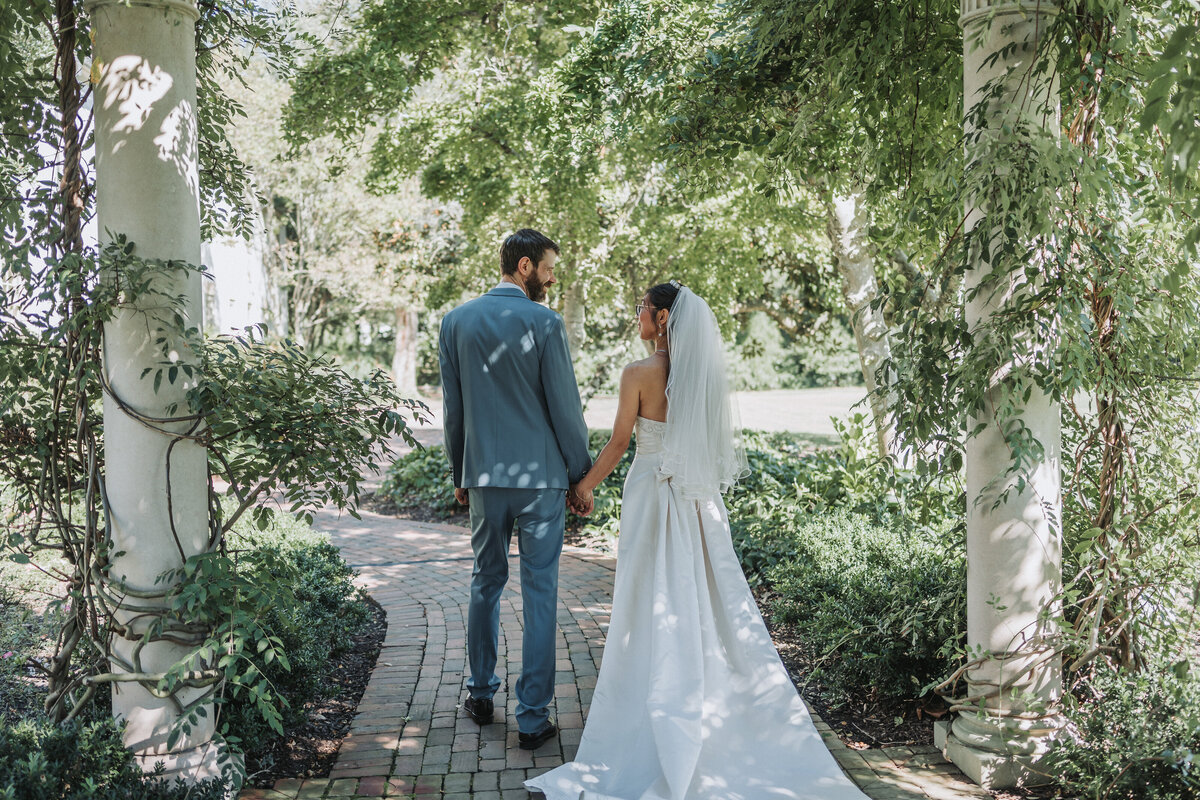 Bride and groom holding hands and walking through the lush gardens of the Colonial Williamsburg Inn. The bride is wearing a white wedding dress and veil, while the groom is dressed in a light blue suit. Shot by Quinn Photography LLC.