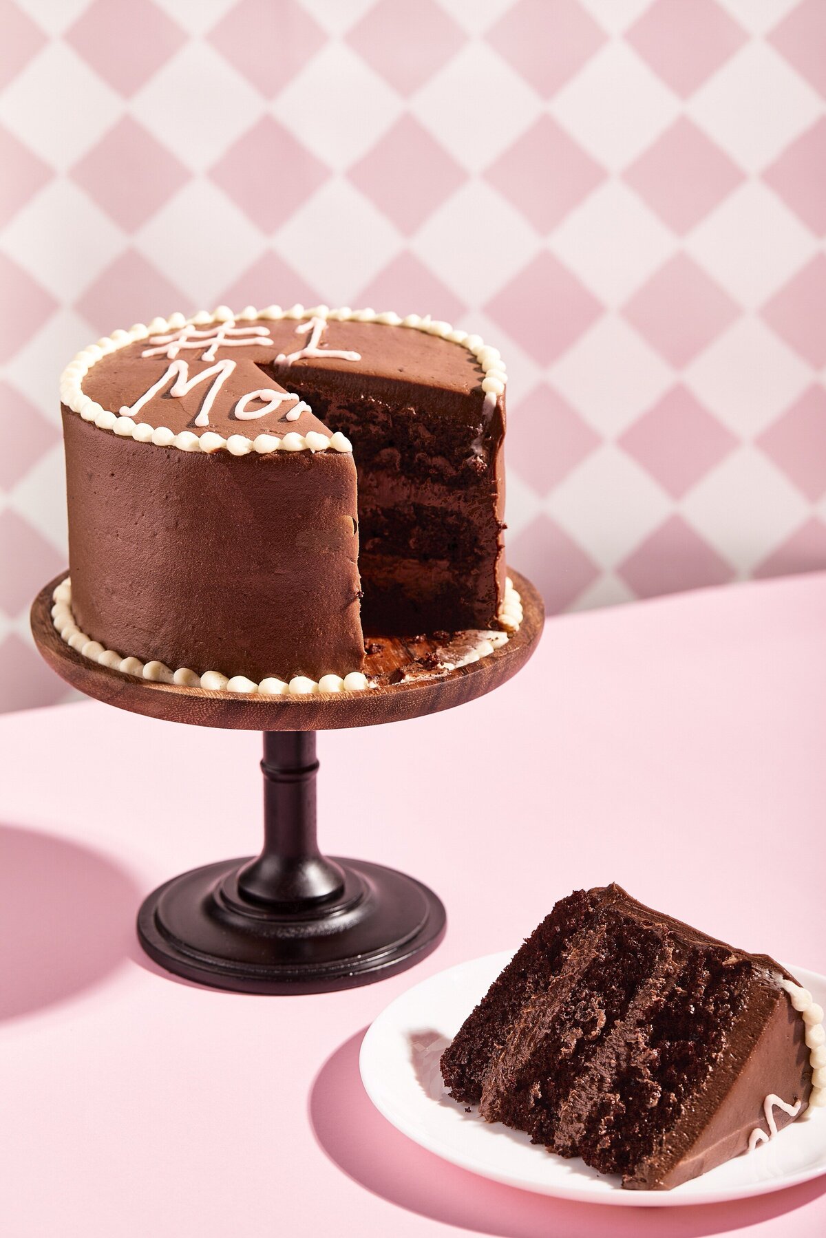 A chocolate cake on a stand with a single piece of cake cut out on a plate below it.