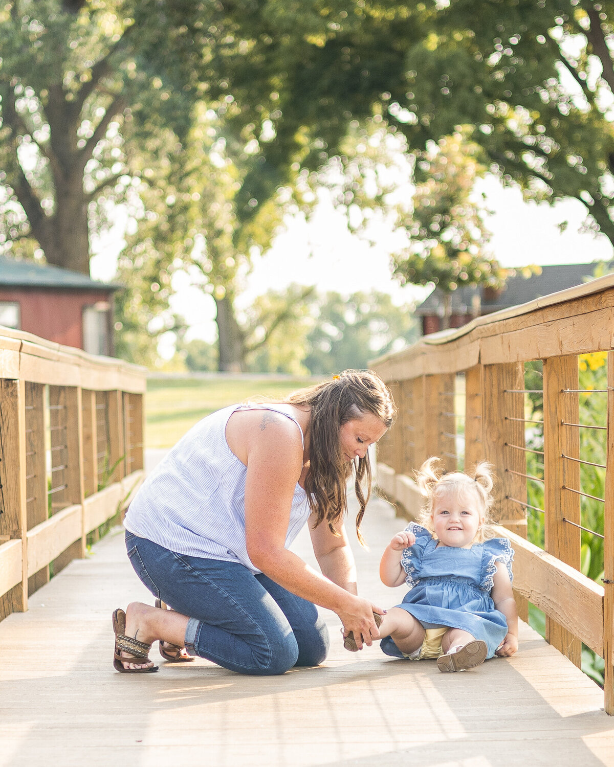 Mom and daughter on a bridge at a wildlife sanctuary.  Mom is putting one of her toddler daughter's white sandals back on and smiling.  Mom has long brown hair, wearing a white sleeveless shirt, blue jeans, brown sandals.  Daughter has blonde pigtails, wearing a blue dress, smiling at the camera