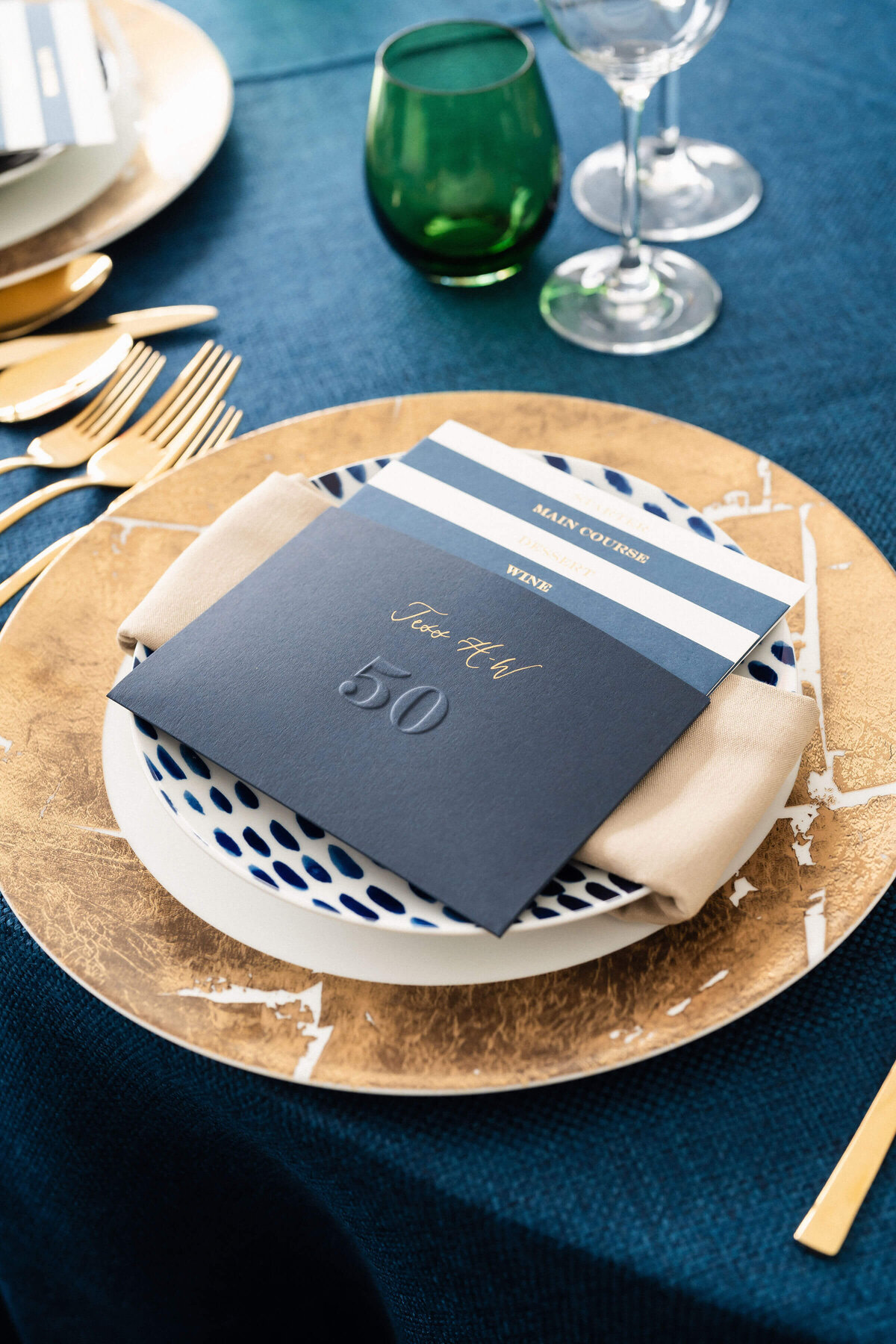 a table setting with a gold charger plate laid on navy blue linen with a navy blue menu on top and green accent water glass next to it for a birthday party
