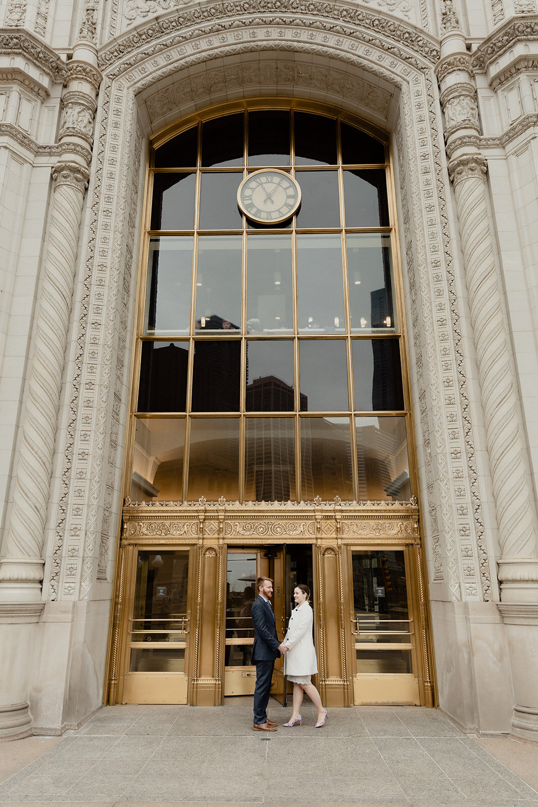 Just Married photo session couple holds hands and looks into each other's eyes in front of large brass doorway at the Chicago Wrigley Building