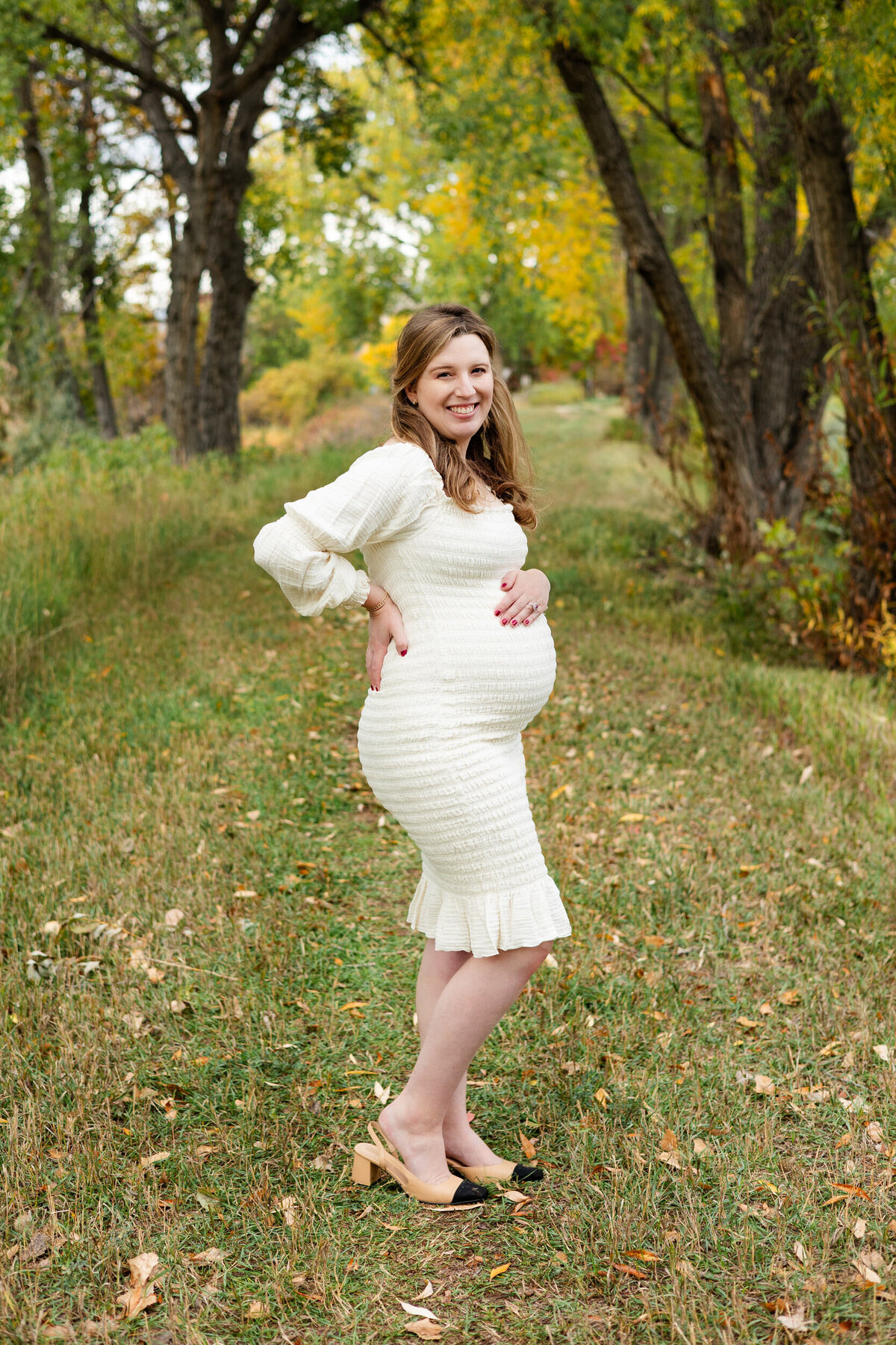 A pregnant woman stands on a wooded path with one hand on her back and one hand on her belly as she smiles at the camera.