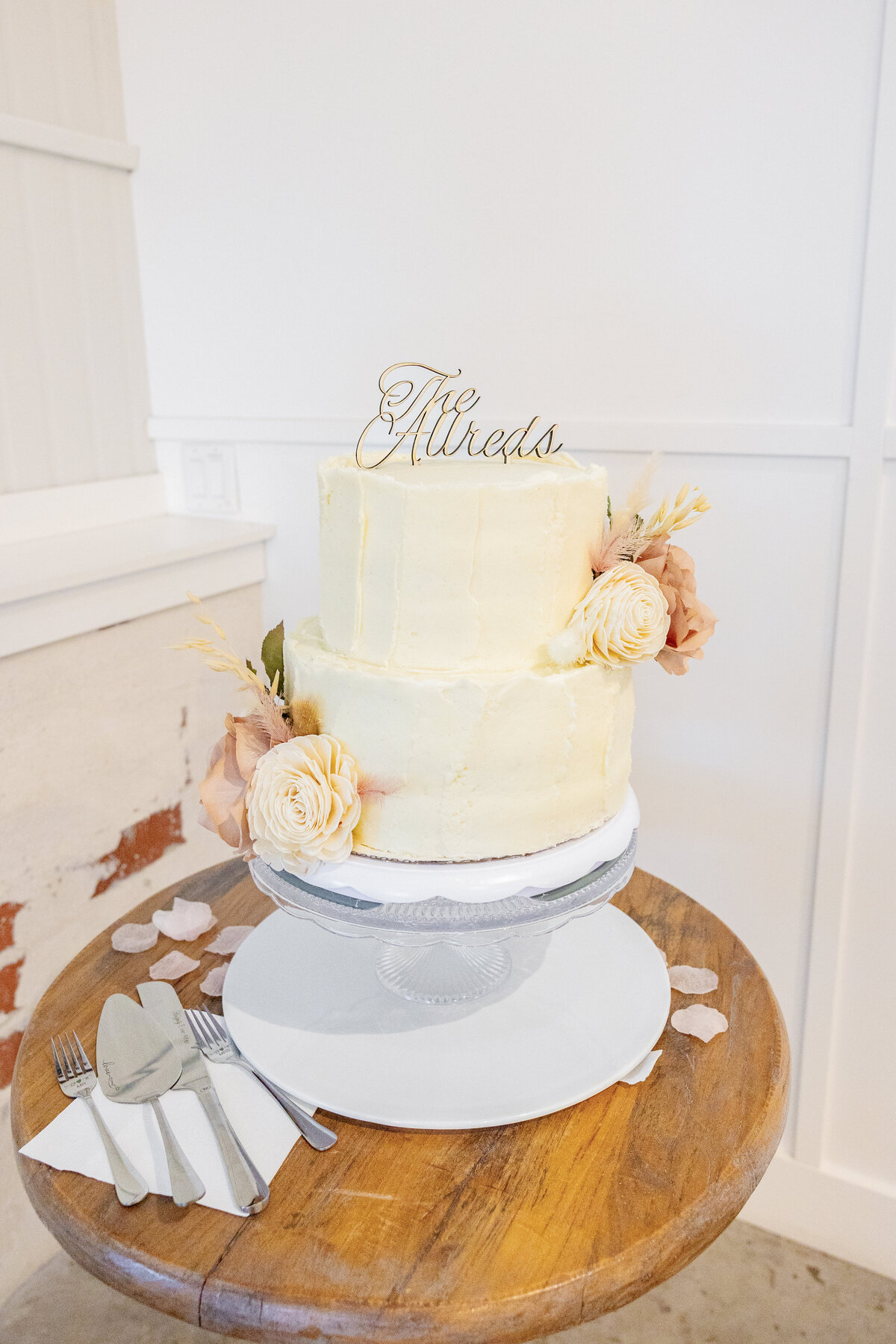 A simple yet elegant two-tier wedding cake with soft buttercream frosting and dried floral decorations, topped with a sign reading "The Allreds."
