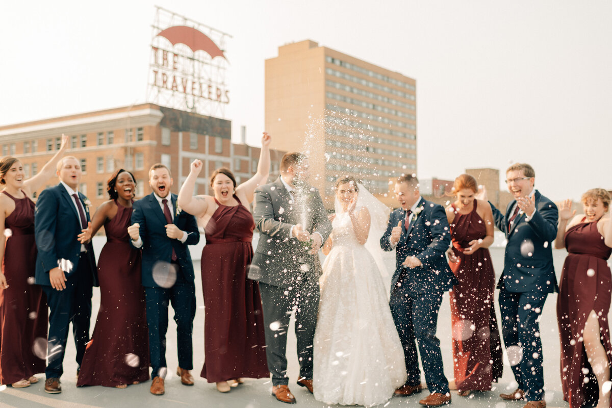 Bride and groom with their bridal party in navy and burgundy spray champagne at their River Center wedding. Des Moines wedding photography by Anna Brace.