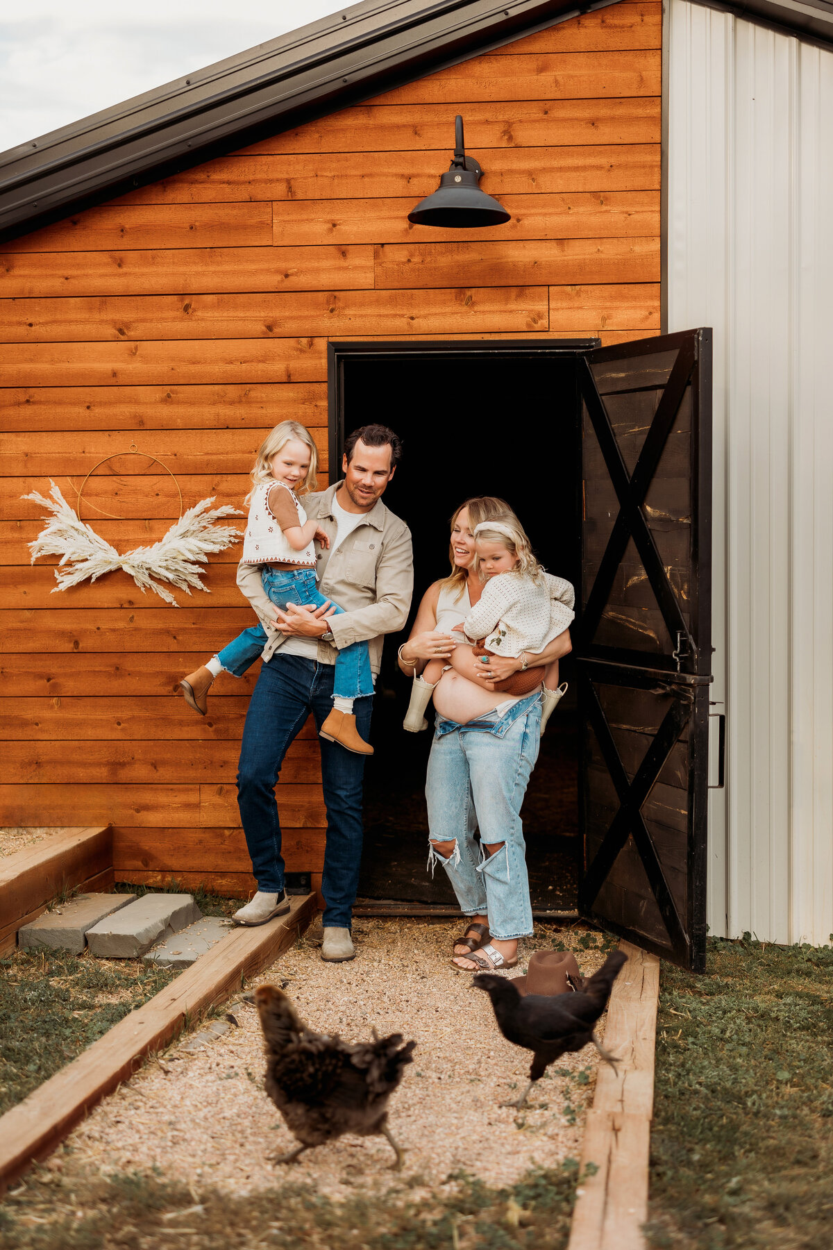 Family of four embrace in a modern barn with two chickens are walking in front of them.