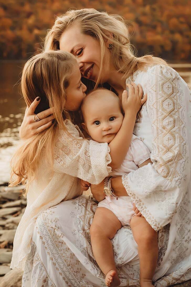 A mother holds her baby on her lap while hugging her older daughter. The older daughter kisses the top of the babys head while the baby looks directly at the camera