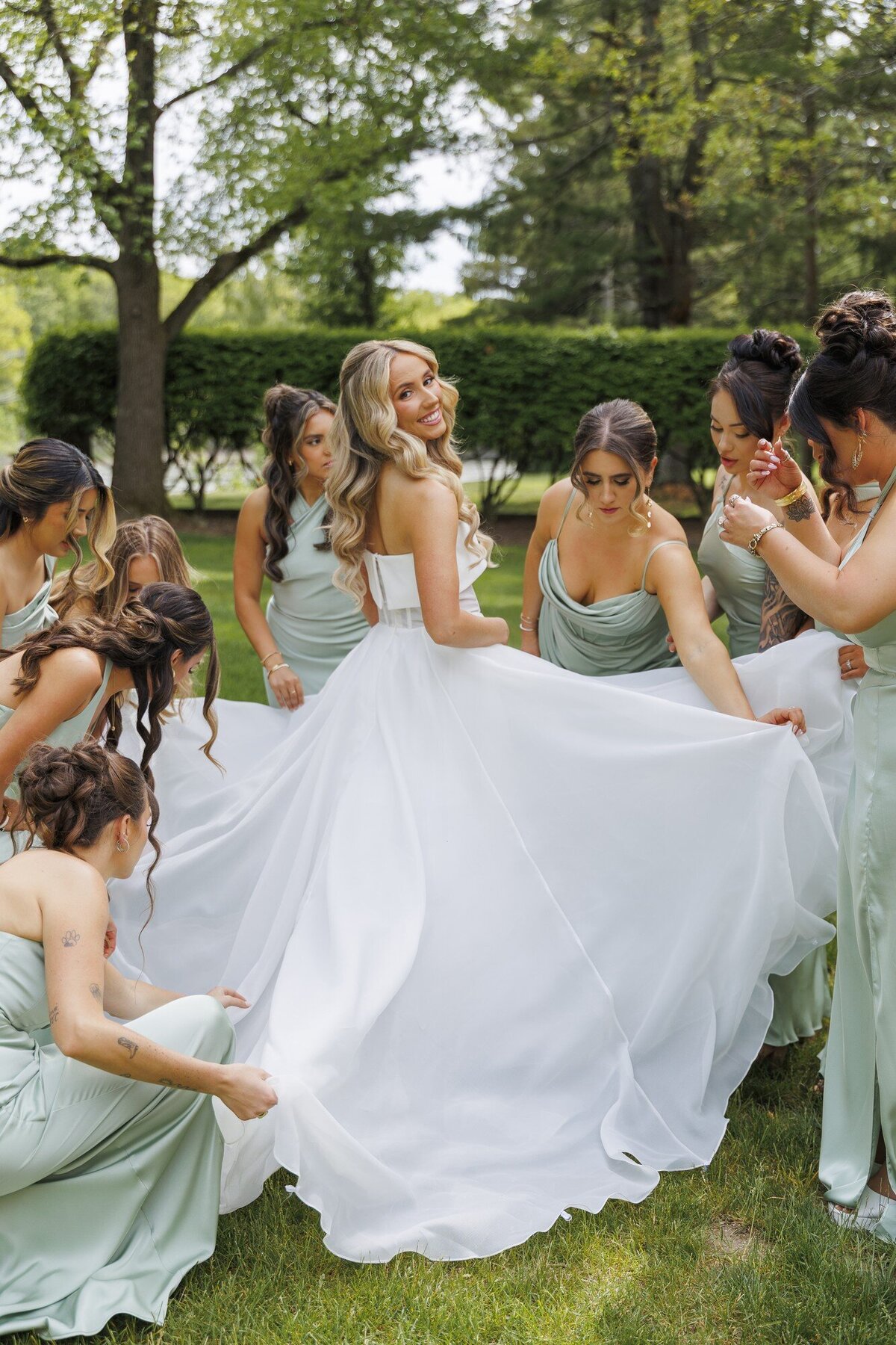 Bridesmaids in sage green gathered around the bride as her gown billows outdoors