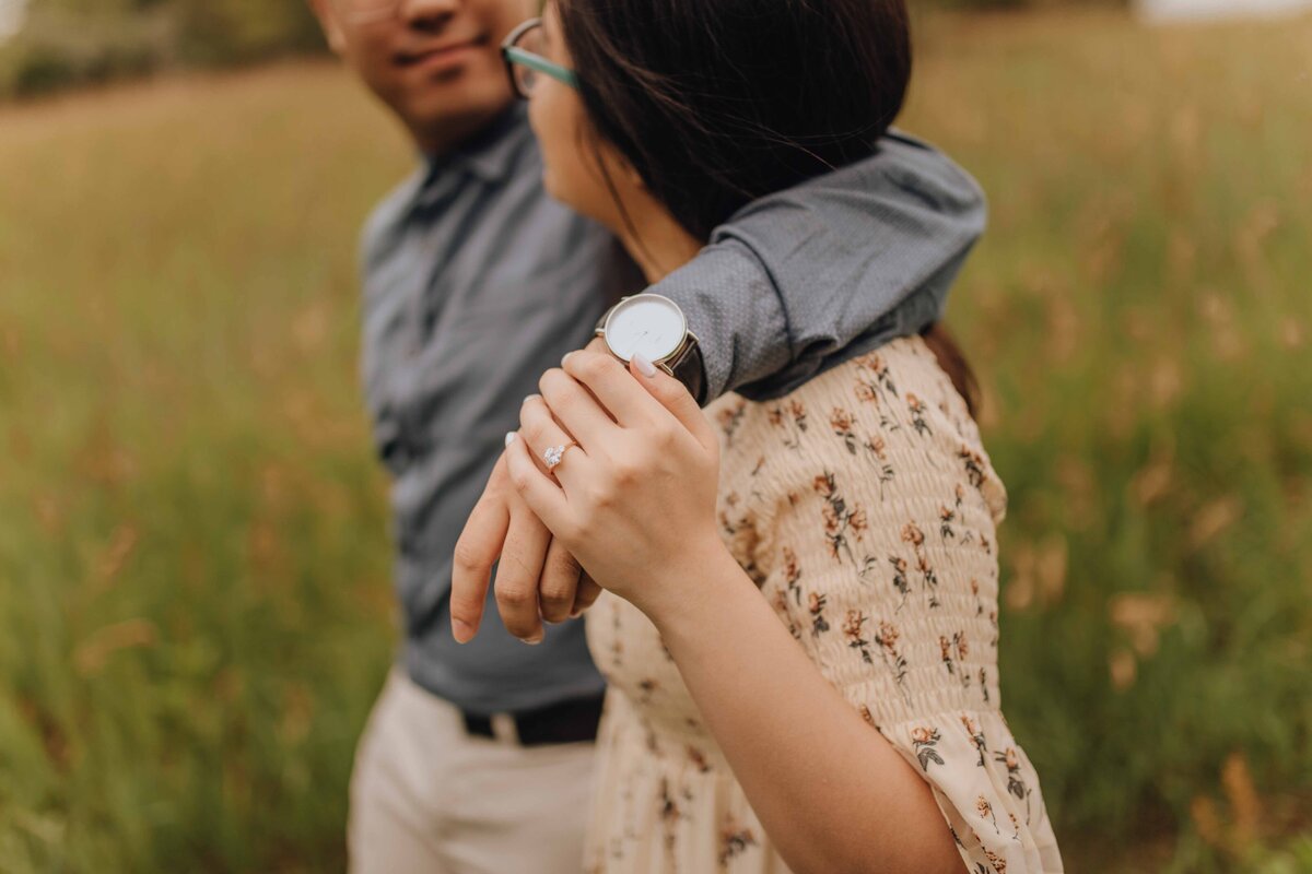 Engaged couple walking through park, man has arm around girl and girl has hand on his wrist. His watch is a close up of the photo as well as her ring.