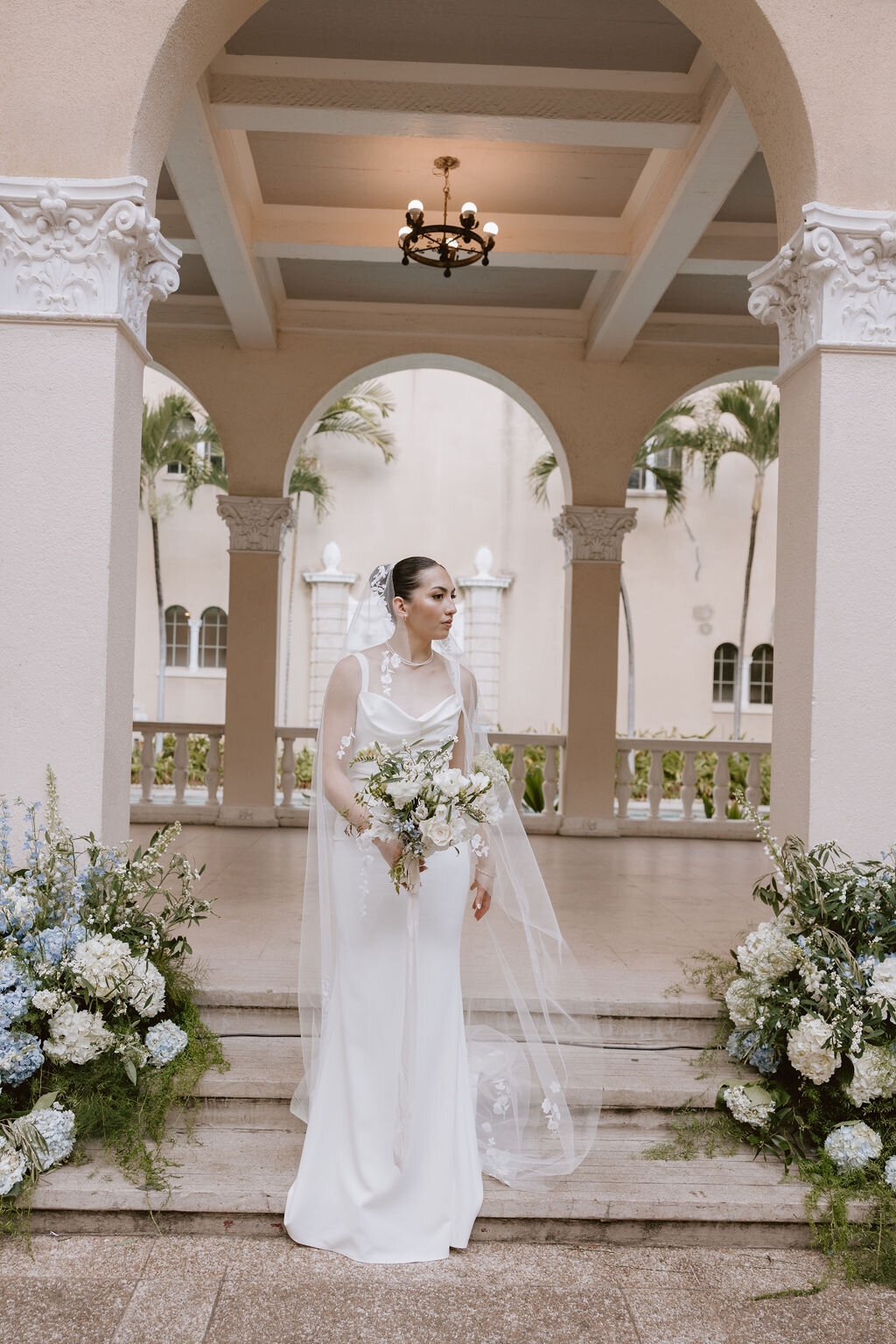bride standing under european arches looking to her side with ground florals on each side of her, holding bouquet