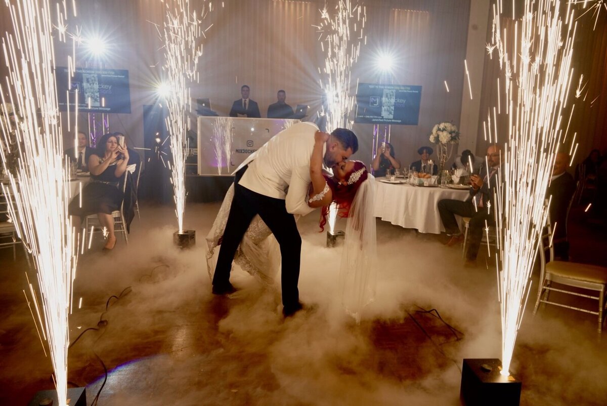 The couple shares a romantic kiss during their first dance, illuminated by the spectacular display of fireworks in the background. The moment captures the magic and celebration of their wedding day, blending the intimacy of their dance with the grandeur of the fireworks show.