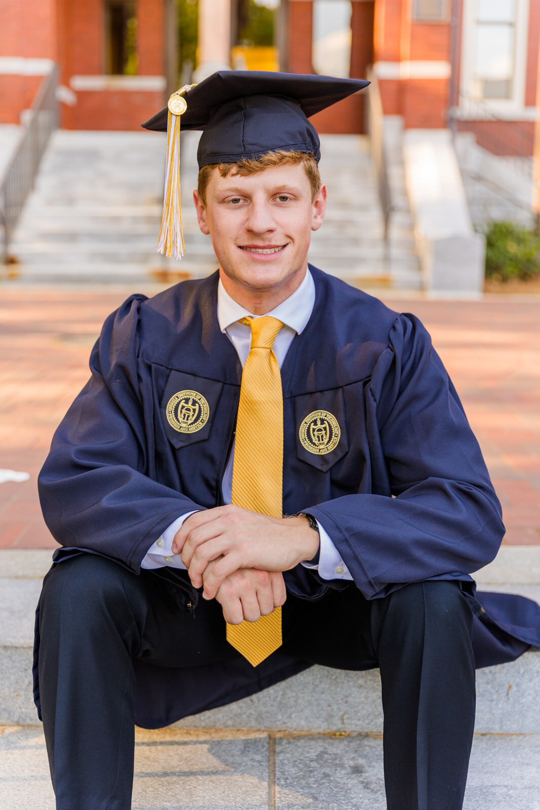 Senior GT boy sitting on stairs in front of Georgia Tech tower wearing cap and gown on top of white shirt and yellow tie facing the camera hands together during portraits on campus by Atlanta Photographer Laure Photography