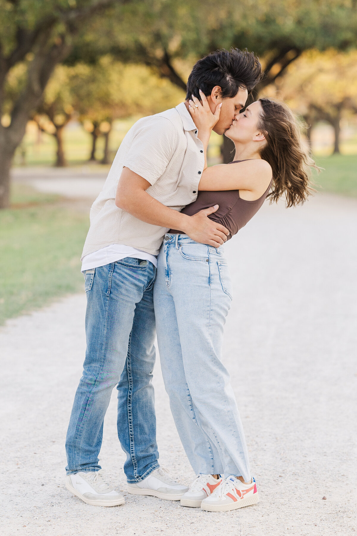 Romantic engagement photo of a fun-loving couple sharing a kiss on a sunlit path in Fort Worth, Texas. Casual outfits and natural surroundings create a relaxed, joyful moment perfect for celebrating their love.