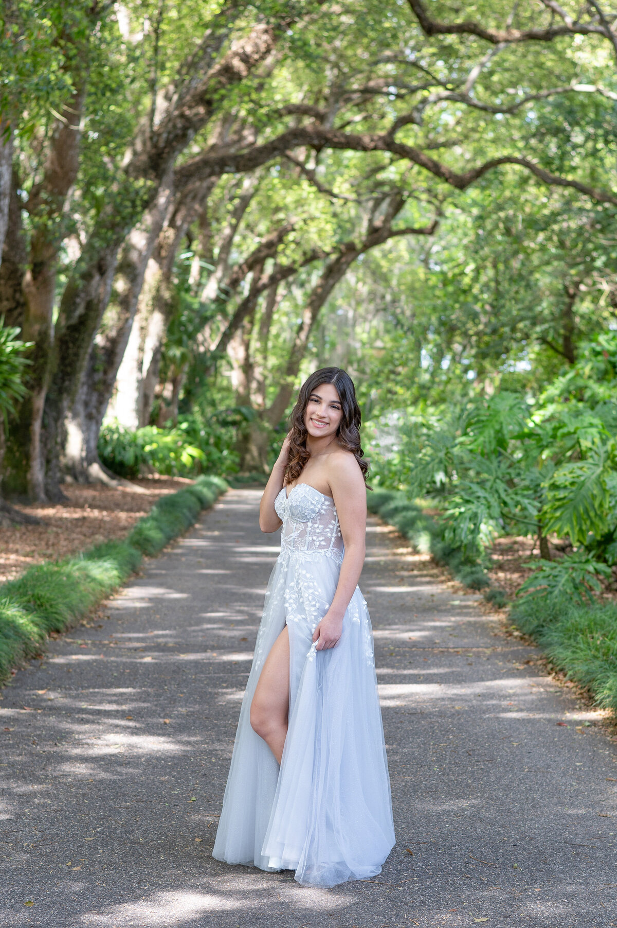 High school senior girl wearing floor length prom dress in middle of path smiles at the camera.