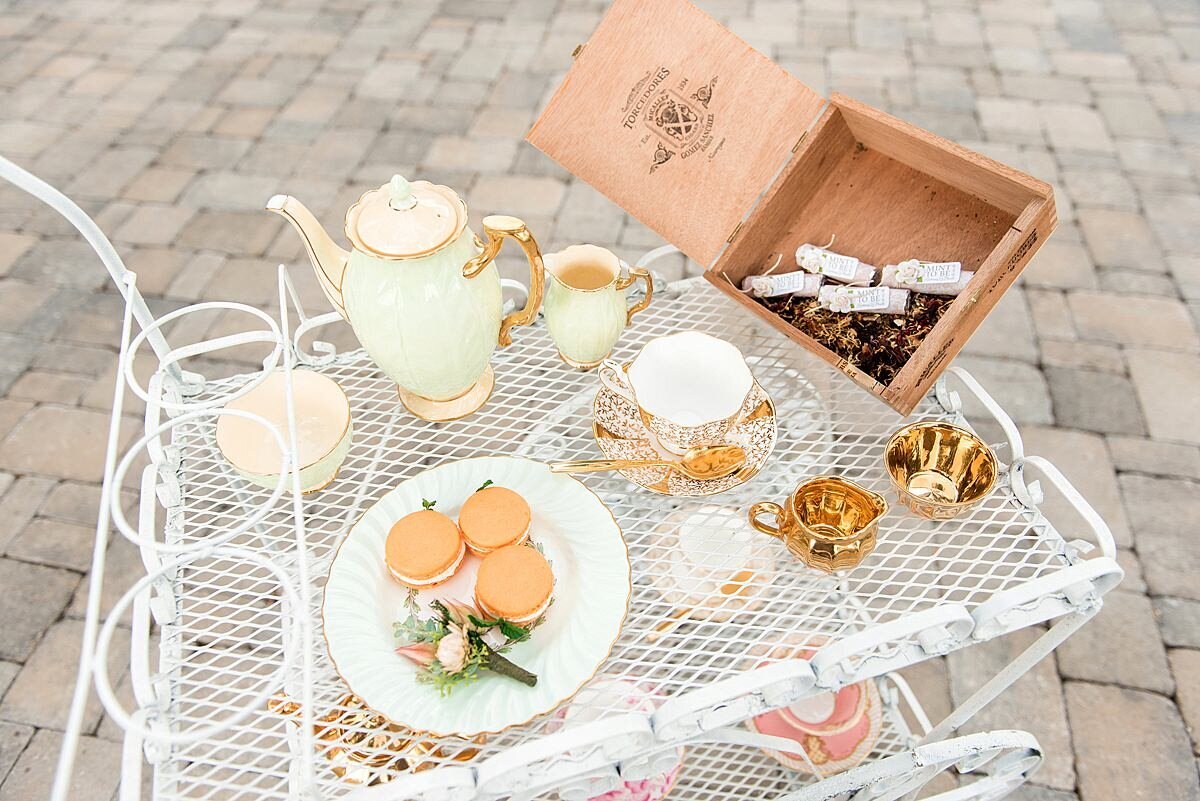 Loose leaf tea and peach macarons sitting on a vintage china dessert plate with the groom's protea and rose boutonniere. The white tea cart has an ivory vintage china tea pot, vintage ivory and cold tea cup and gold cream and sugar bowls.