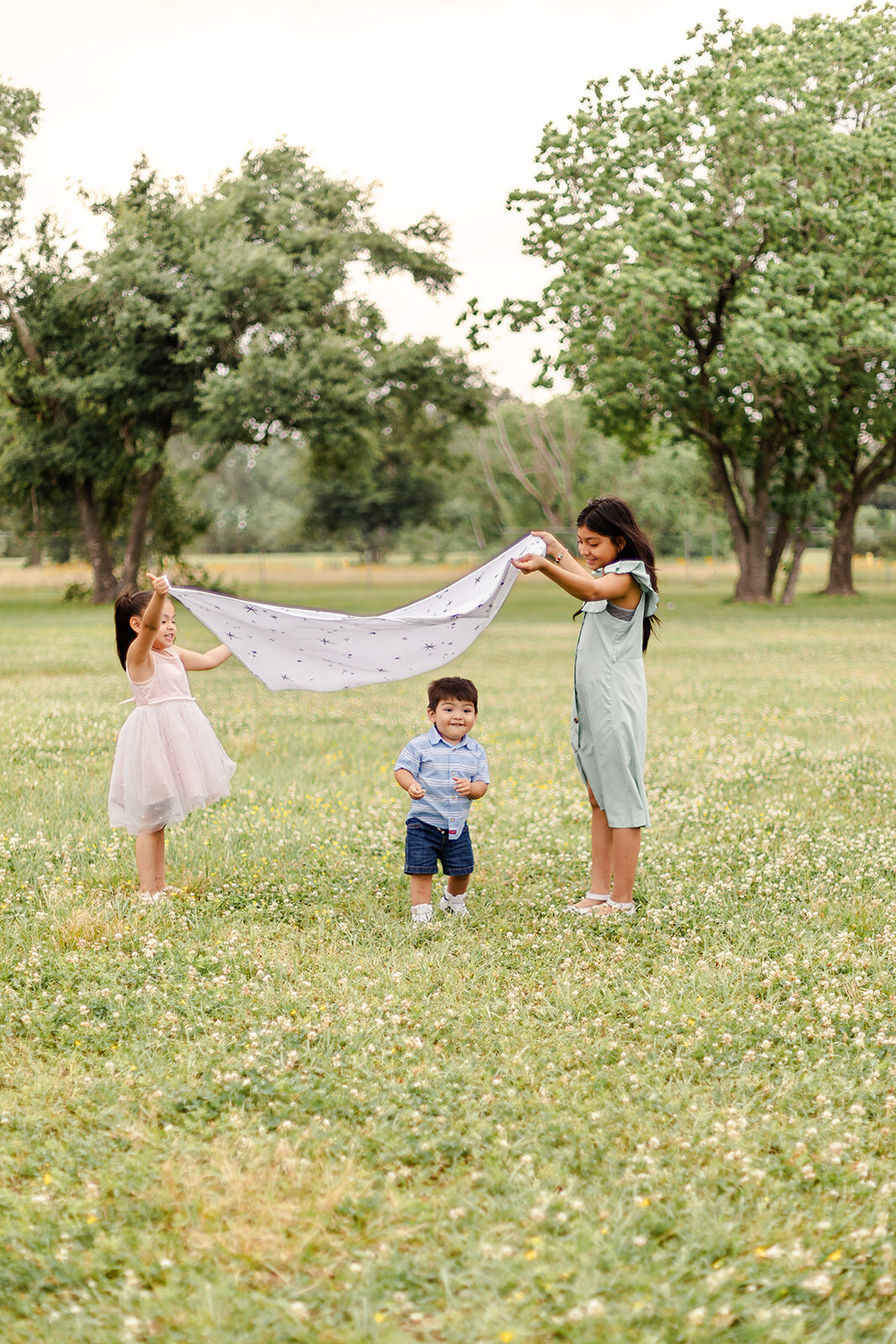 three children playing in a grassy field on a cloudy day. Two girls are holding up a light, white blanket with a subtle pattern, creating a canopy over a younger boy standing between them. The girl on the left is wearing a light pink dress, and the girl on the right is wearing a light green dress. The boy in the middle is wearing a blue striped shirt and dark shorts. They are surrounded by green grass and clover flowers, with tall trees in the background, giving the scene a peaceful, outdoor setting.