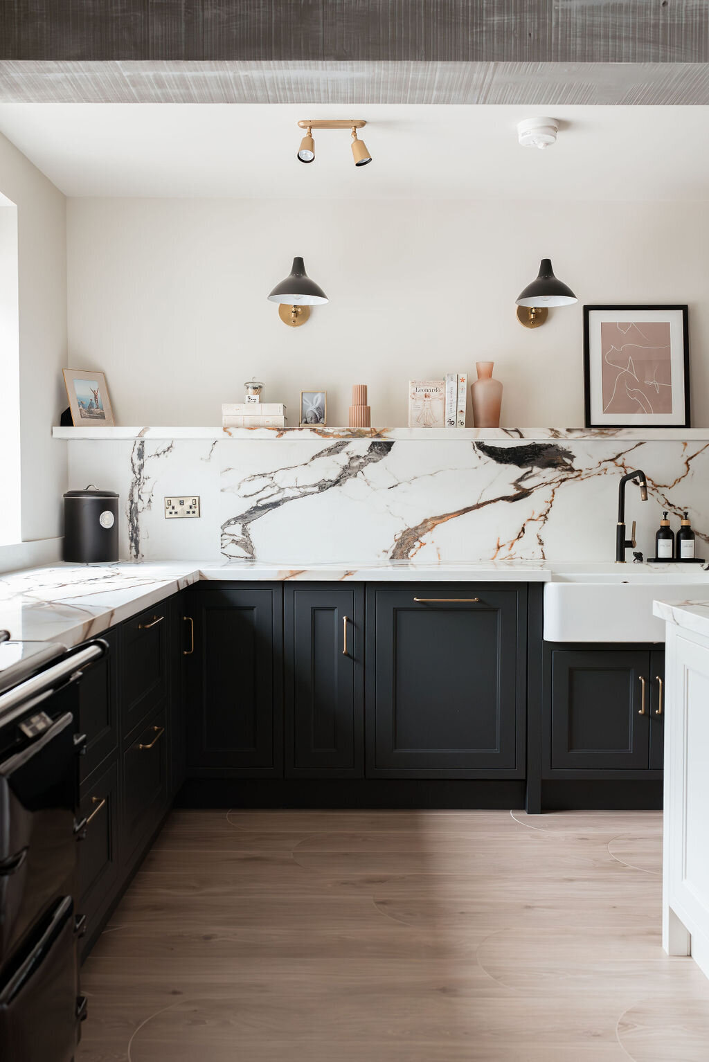 A modern kitchen with dark lower cabinets, a white marble backsplash with black and gold veining, a farmhouse sink, and minimalist open shelving holding books and decor. Two wall-mounted lights illuminate the space, which also features light wood flooring.