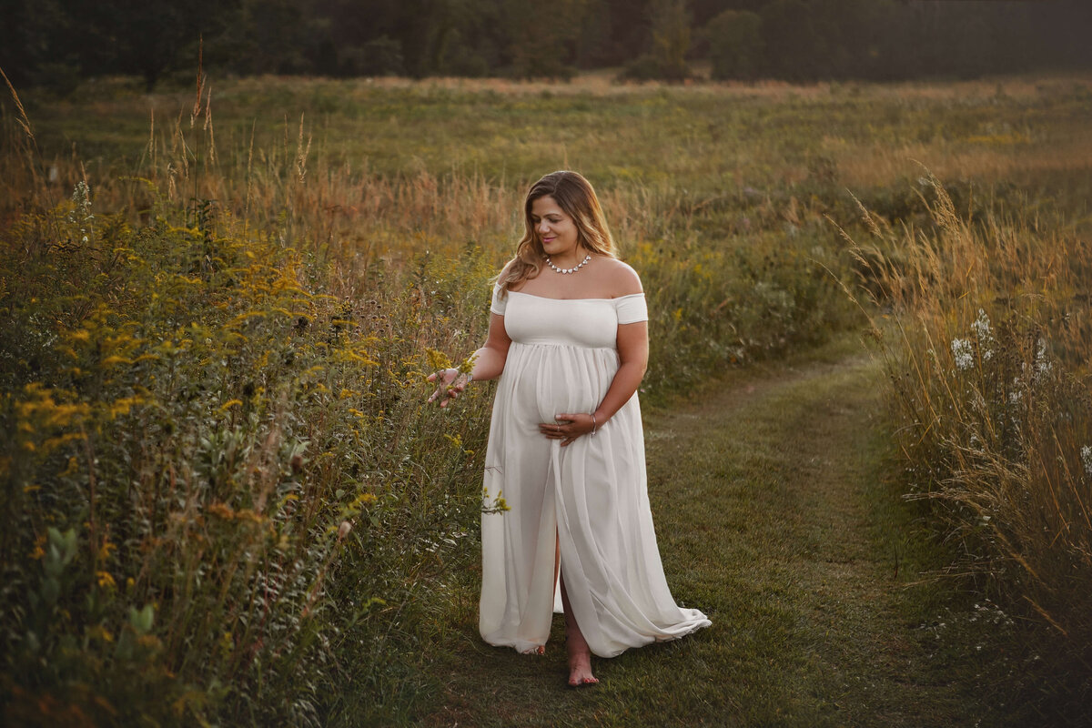 pegnant mothe wearing a cream maternity gown walking down a path between flowers at sunset in the summer