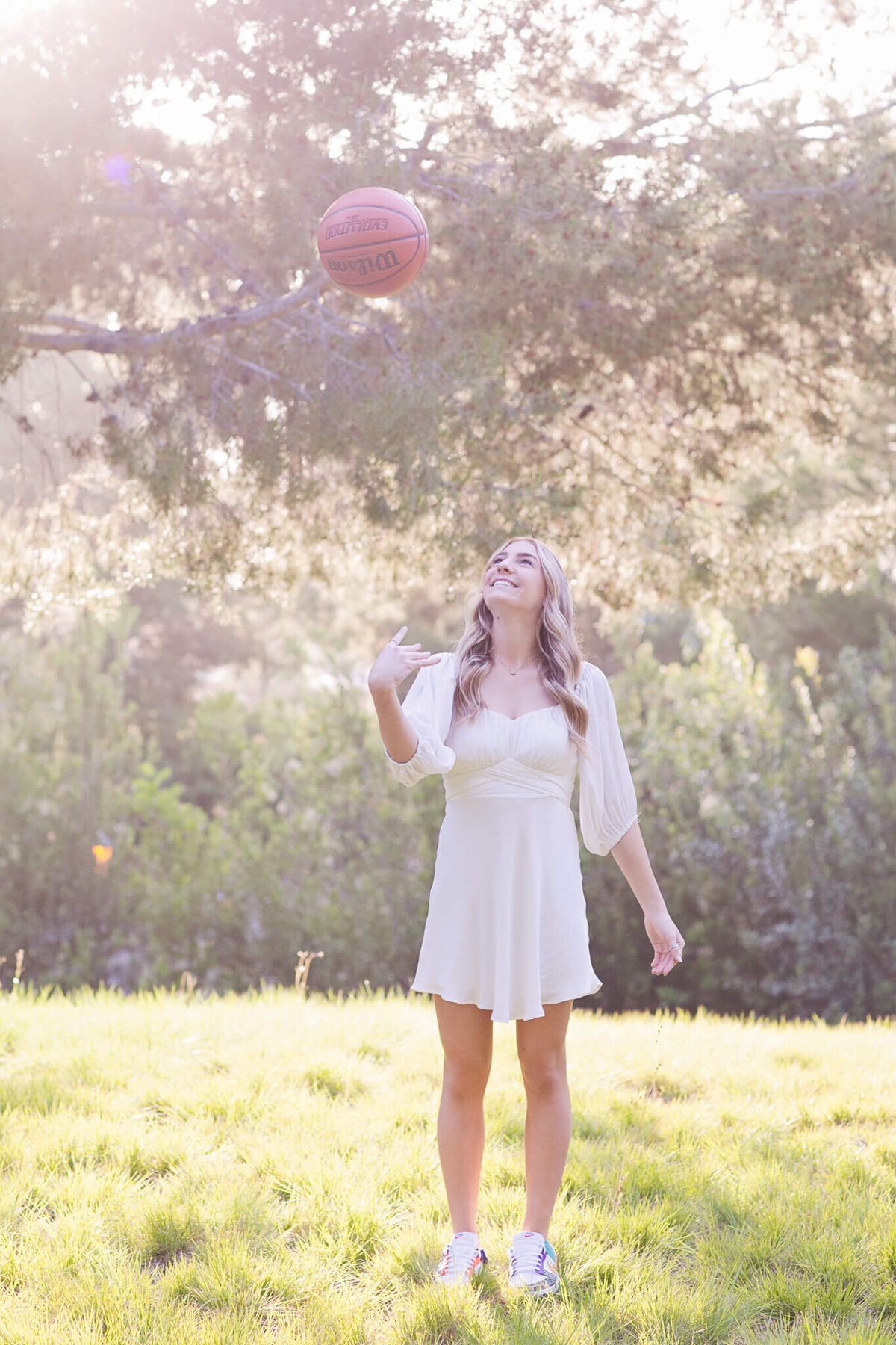 A young woman in a white dress and sneakers joyfully tosses a basketball into the air. She stands on a lush green lawn with trees in the background, bathed in soft, warm sunlight.