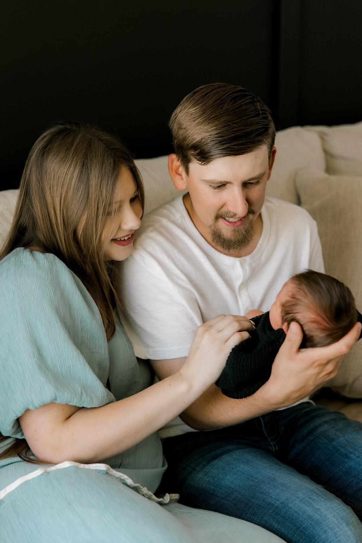 Young couple sitting on a cream-colored sectional couch, lovingly cradling their newborn baby in a cozy, natural light setting.