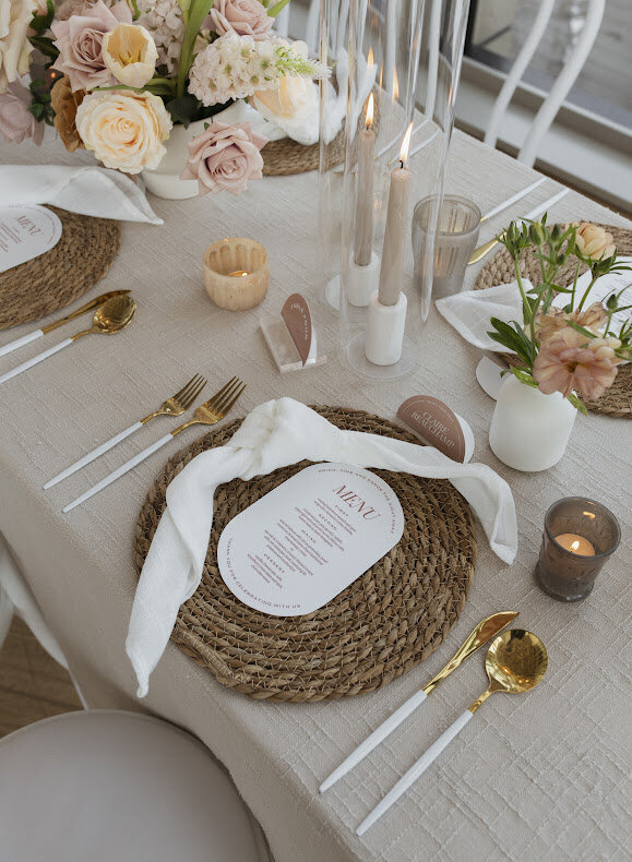 A close-up of a bohemian guest table at a National Arts Centre wedding, featuring beige textured linens with a series of taupe tapered candles surrounded by white bud vases with peach, cinnamon and white fresh flower booms and a modern half-circle table number. Each place setting has a knotted white linen napkin and an oval menu on top of a woven rattan charger plate with gold and white cutlery on each side.