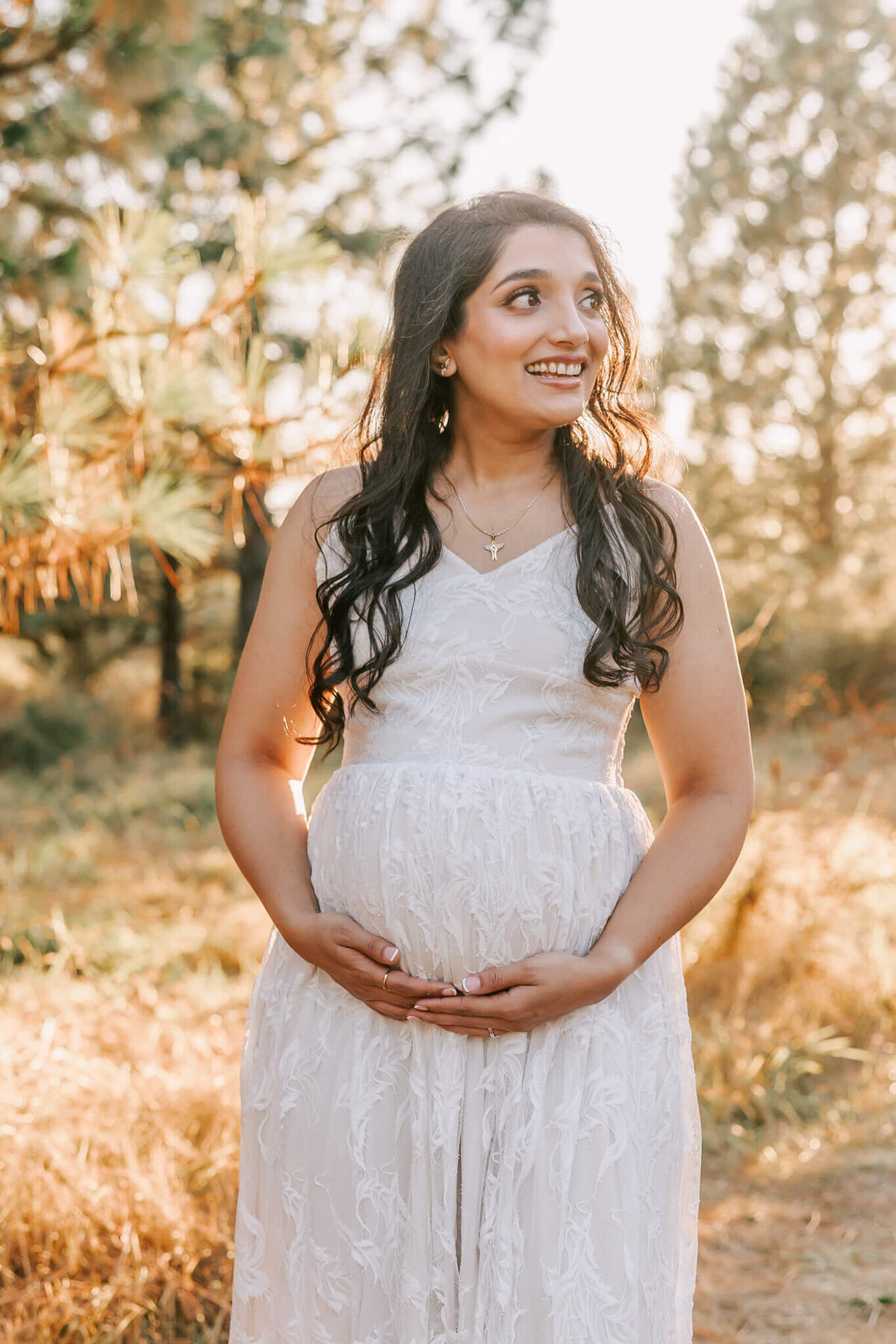 Portrait of woman with long brown hair smiling to right and walking towards camera with her hands under her belly in pine trees.