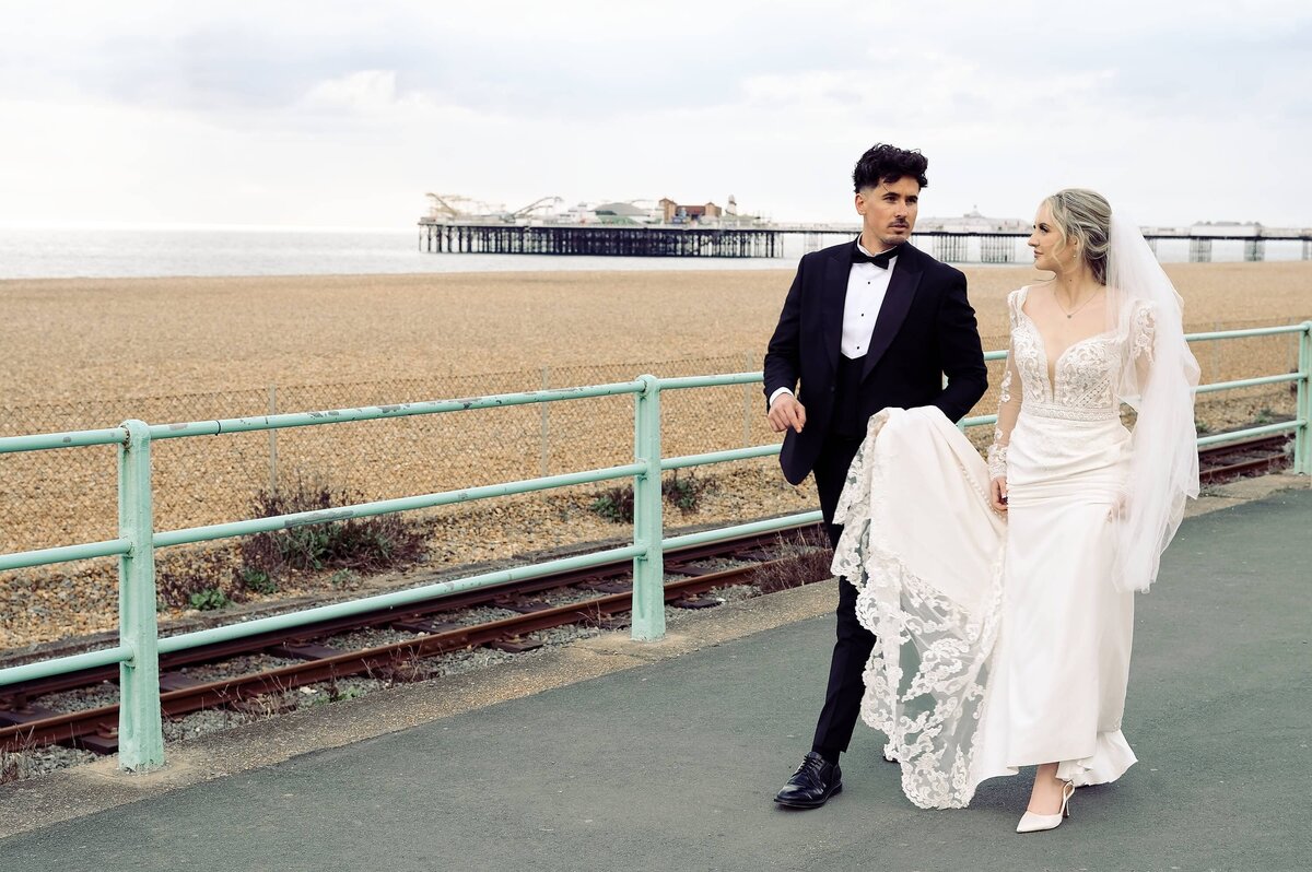 Bride and groom walking along the seafront at Brighton with a view of the pier and beach in the background