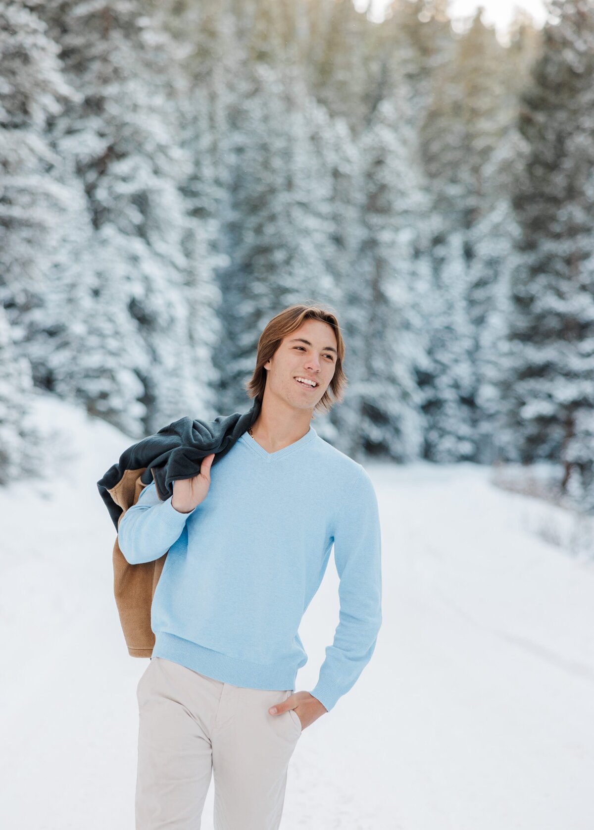 A teen boy in a blue shirt walks in snow with jacket over his shoulder