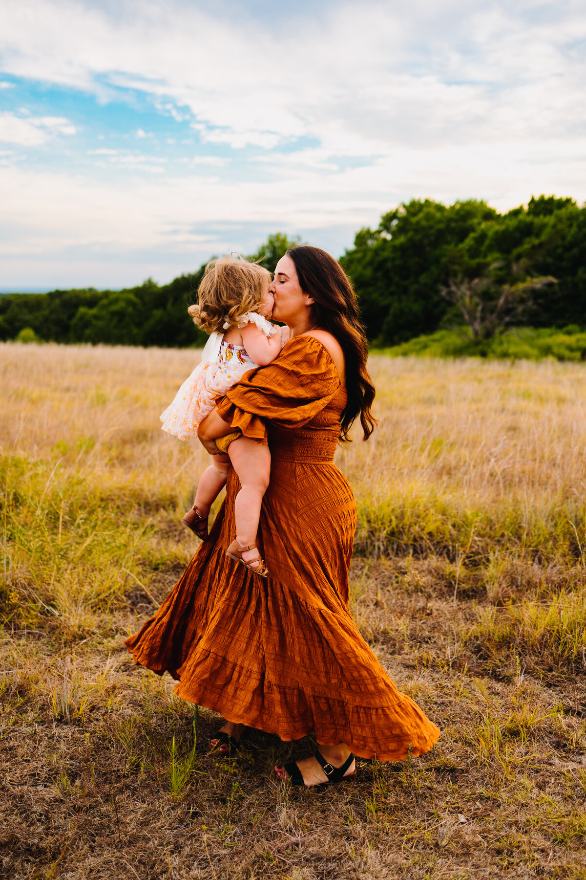 Maternity and family photography packages in Albuquerque feature a beautiful scene of a mother wearing an orange vintage long dress, carrying her little daughter. The setting is a serene forest with lush green trees