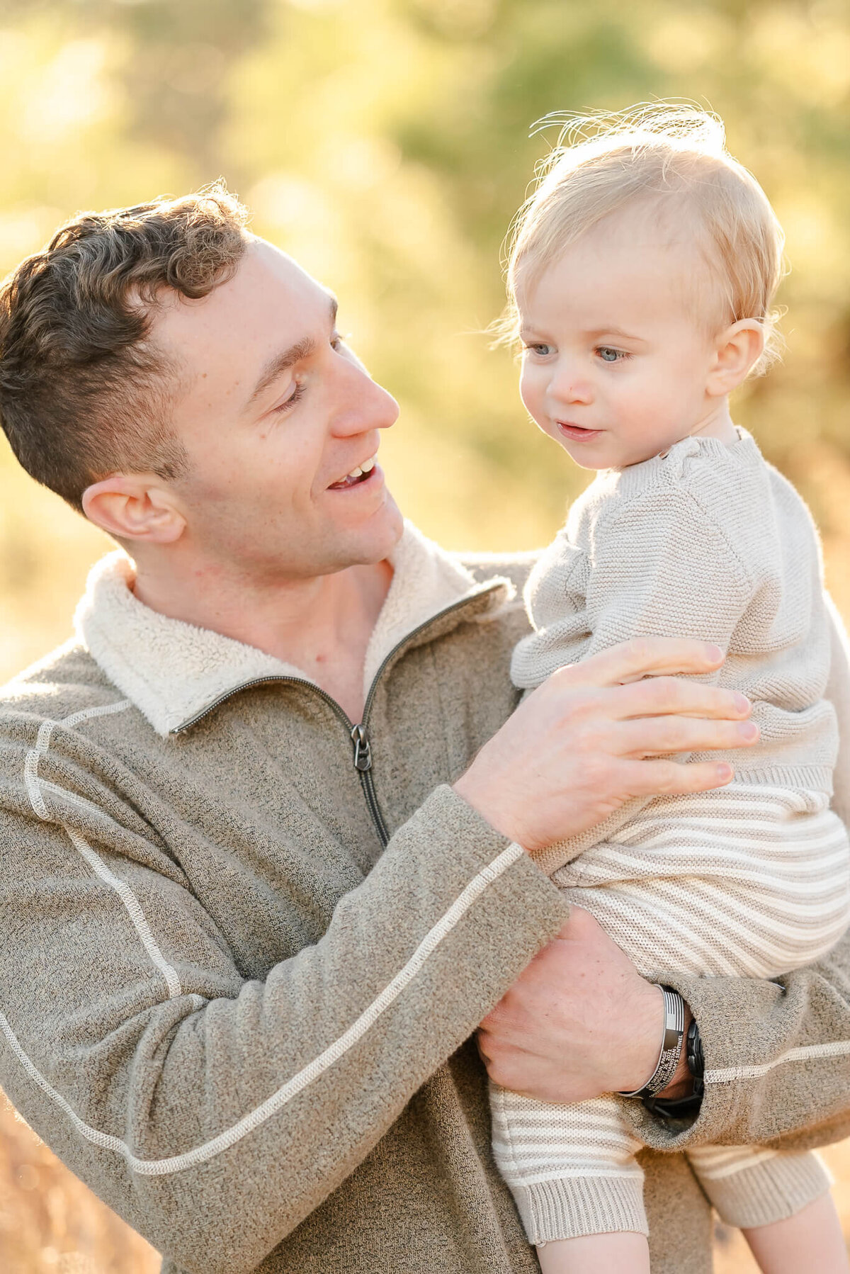 A man, wearing a neutral sweater, holds his toddler son, who is also wearing neutrals during a family photoshoot.