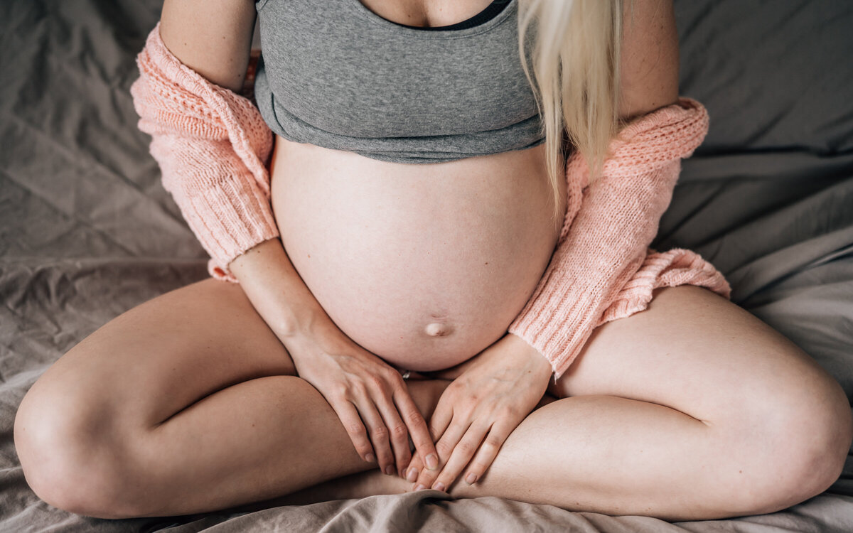 A pregnant mama sits cross legged on her bed with a pink sweater sliding off her shoulders and her baby bump on gorgeous display.