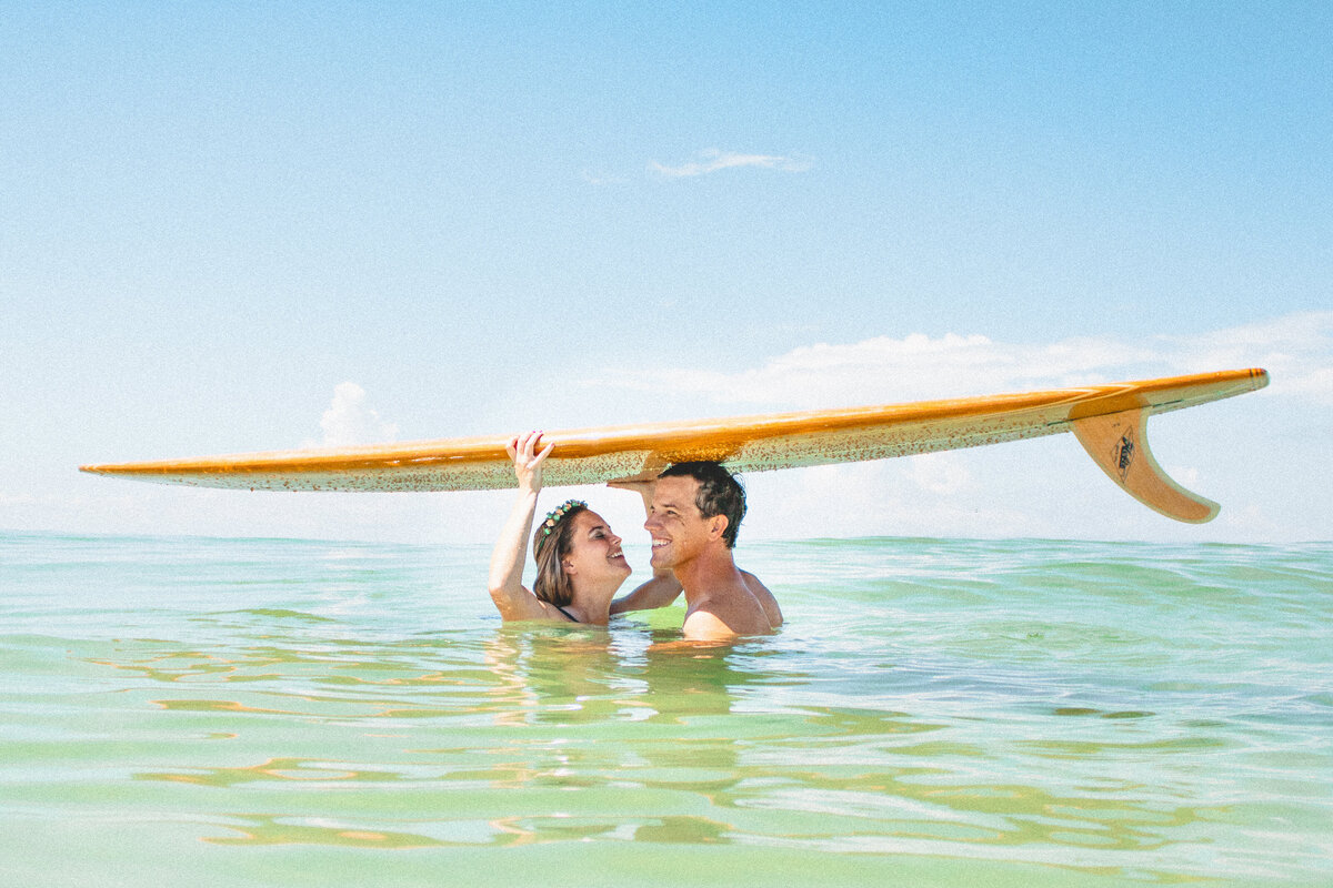 A couple holds a longboard overhead while smiling at each other in the ocean