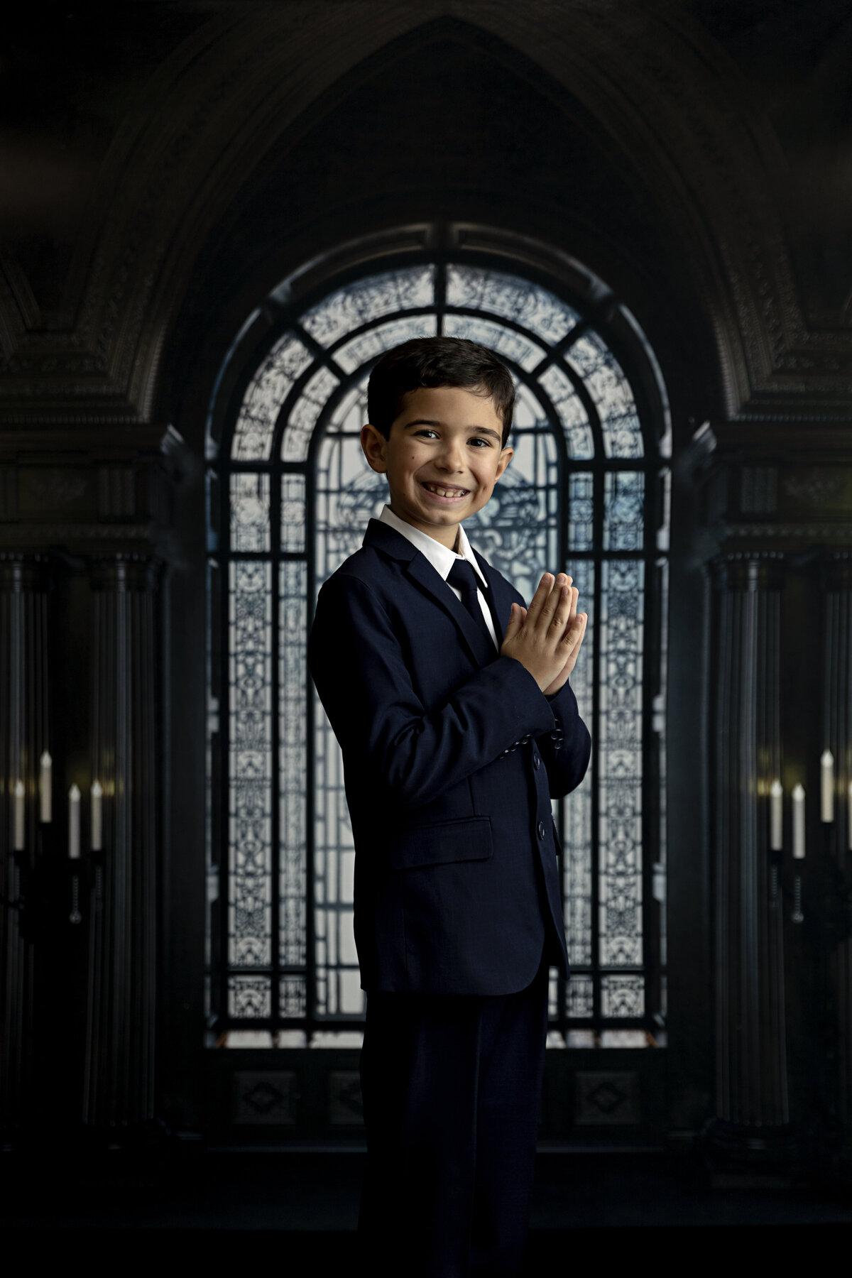 A young boy in a dark suit stands in a church smiling and praying