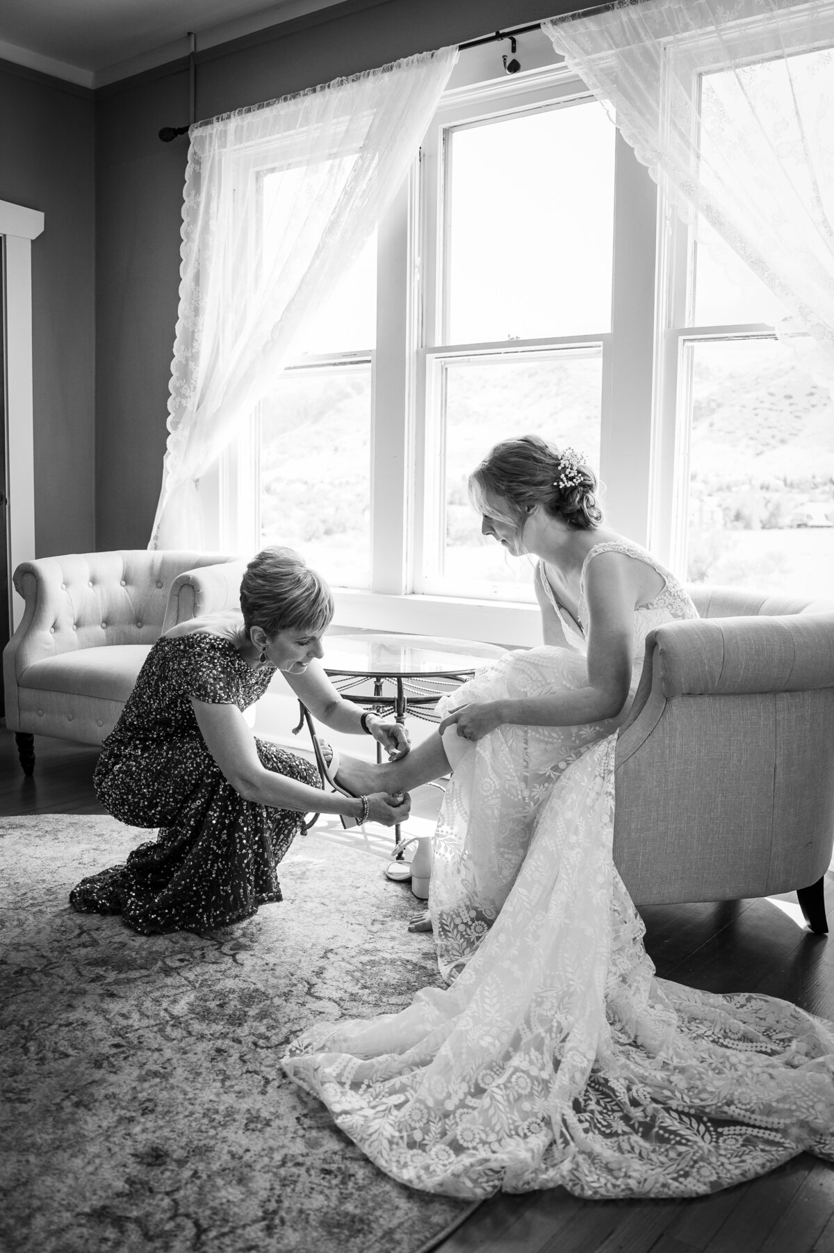 A bride sits on a couch as her mother helps her put her shoes on.