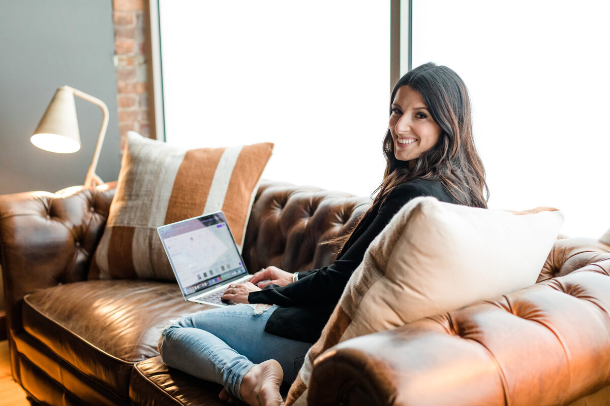 Allie Clark sitting on a vintage leather couch with her MacBook open to a design platform, smiling warmly — reflecting the relaxed yet refined vibe of Allie Clark Designs.