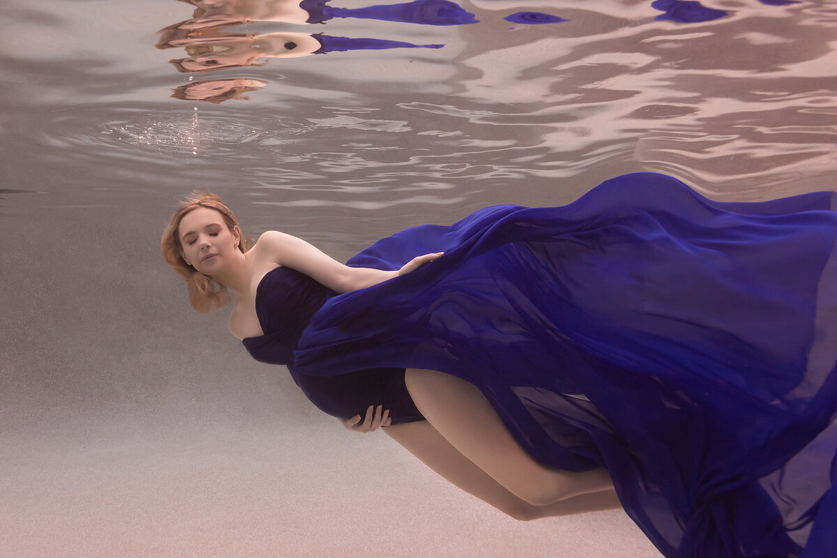 A serene underwater maternity photo captured by a talented Dallas underwater pregnancy photographer. The woman is gracefully floating underwater, her deep blue gown billowing around her, creating a mesmerizing, flowing effect. Her eyes are gently closed, exuding a sense of peace and calmness. This underwater maternity photoshoot highlights the elegance and beauty of expectant motherhood with a soft, ethereal quality.