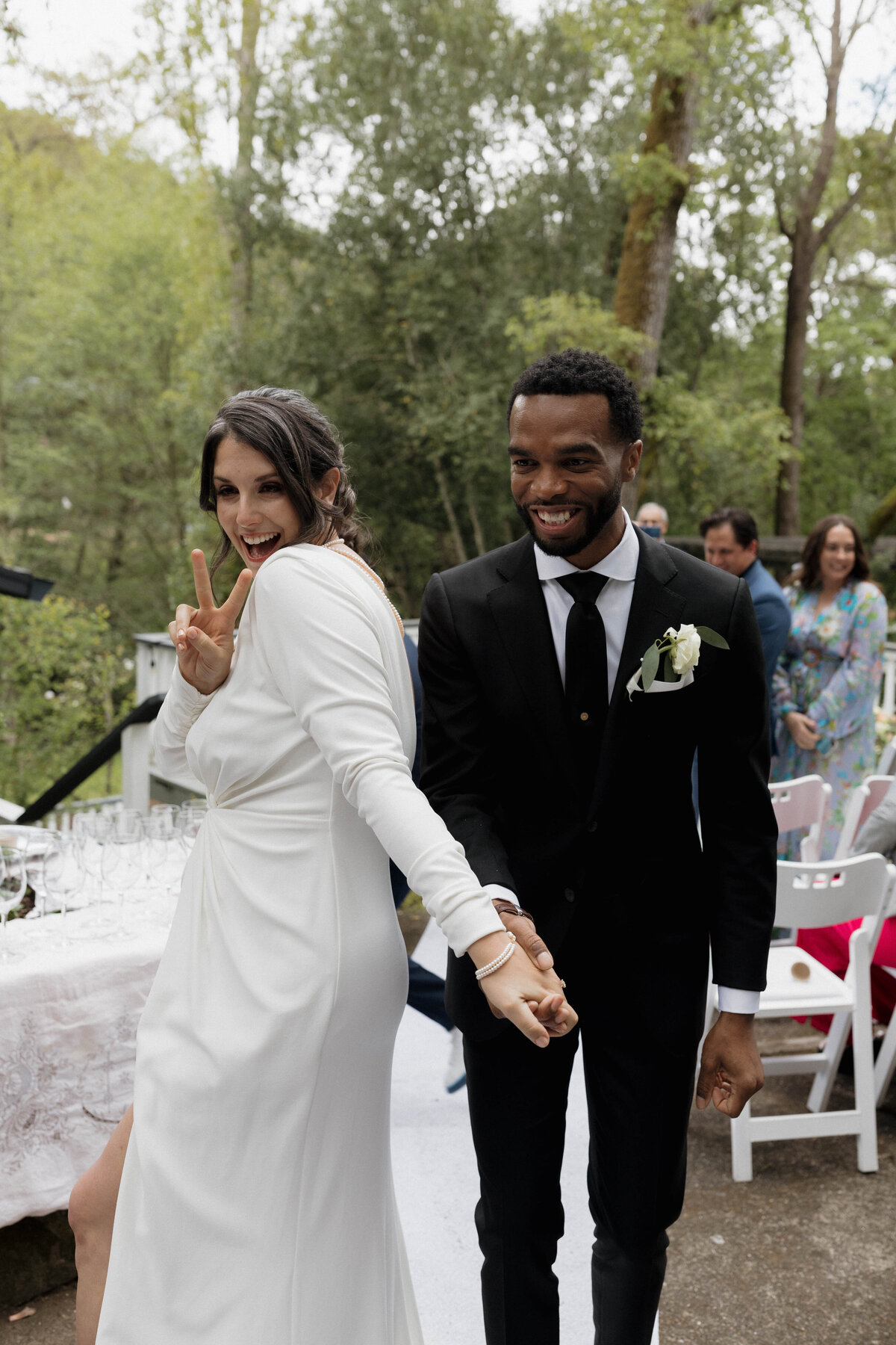 A playful, candid moment capturing a bride and groom happily exiting their outdoor ceremony. The bride gives a peace sign while holding the groom’s hand. Captured by a luxury wedding photographer specializing in authentic, cinematic storytelling imagery.