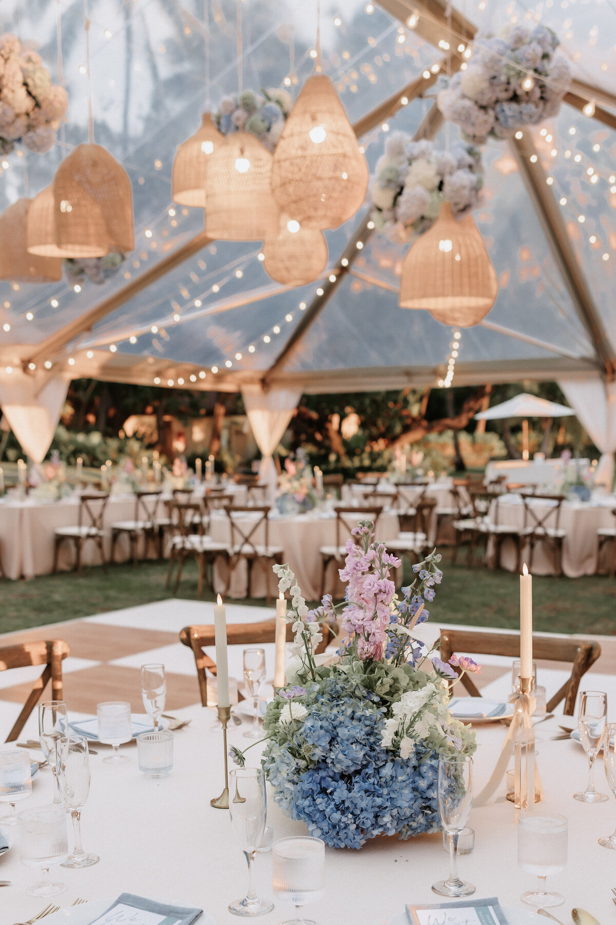 Wedding reception at lanikuhonua under a tent with hanging lantern installation and a mixture of round and rectangle tables with ivory tableclothes, and blue purple white and green floral centerpieces.