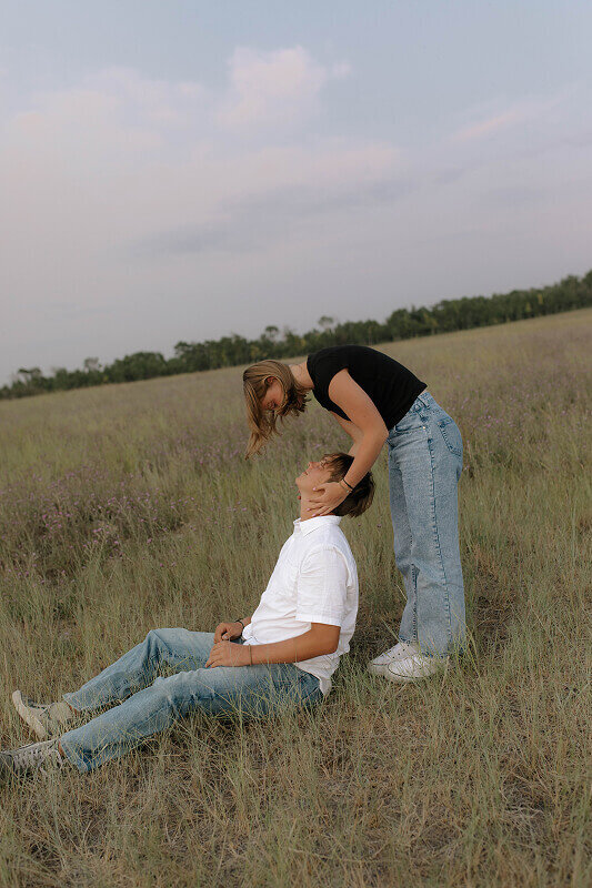 A woman holds her boyfriend's face from behind as he's sitting on the ground during their couples engagement photos.