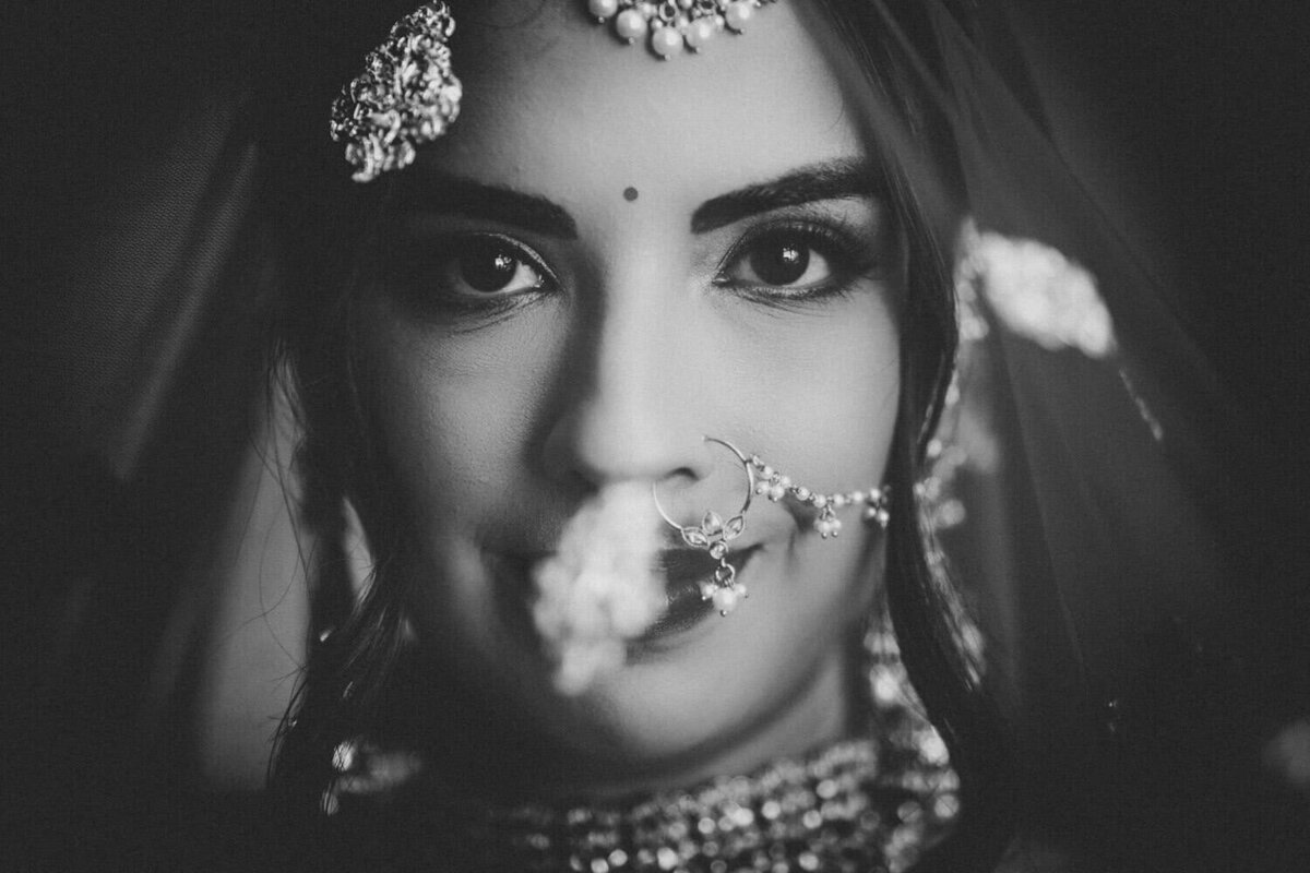 A striking black and white close-up portrait of a bride adorned in traditional jewelry. Her expressive eyes and delicate nose ring with intricate details stand out, framed by a softly blurred veil. The composition captures an intimate and timeless moment, highlighting her elegance and cultural heritage.