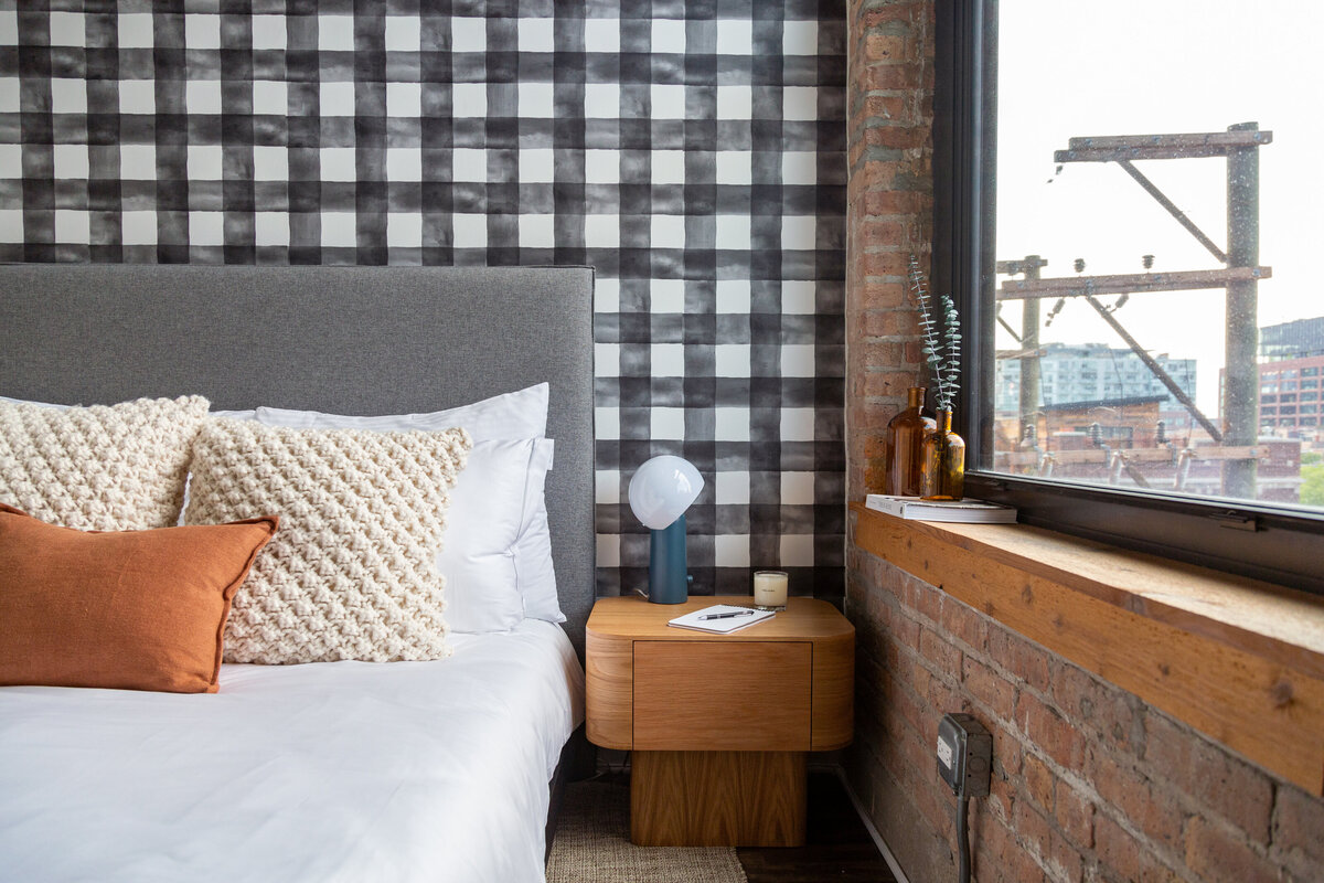 Black and white Buffalo check wallpapered bedroom with grey fabric headboard, white knitted pillows, rust orange linen lumbar pillow, wooden pedestal single drawer nightstand next to brick wall with wooden window sill