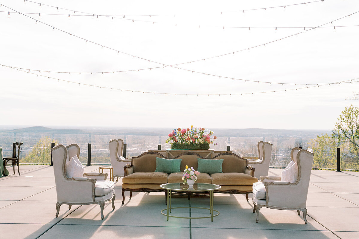 Outdoor lounge seating at Burritt on the Mountain during Callie and Hayes’ wedding, featuring a vintage brown sofa, ivory armchairs, floral arrangements, and string lights with panoramic Huntsville views.