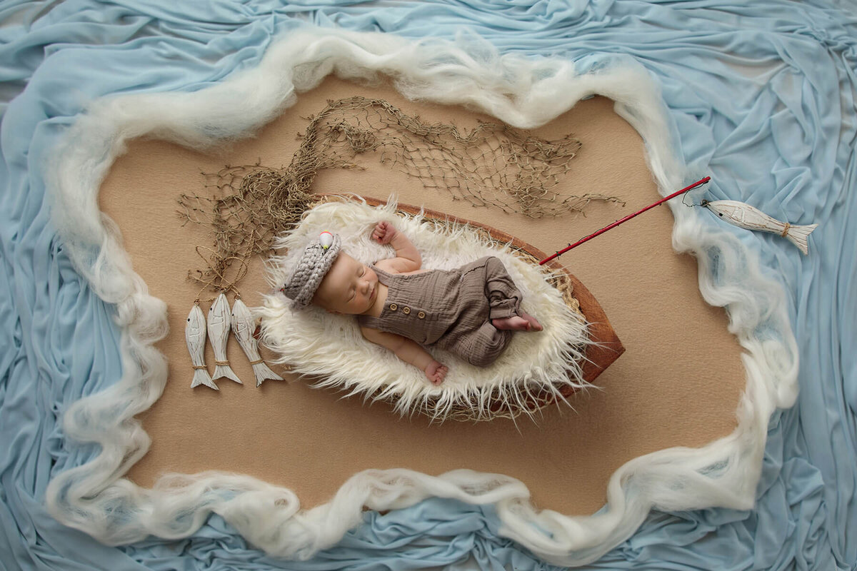 Newborn baby boy sleeps in a wooden boat prop, holding a fishing pole surrounded by faux water.