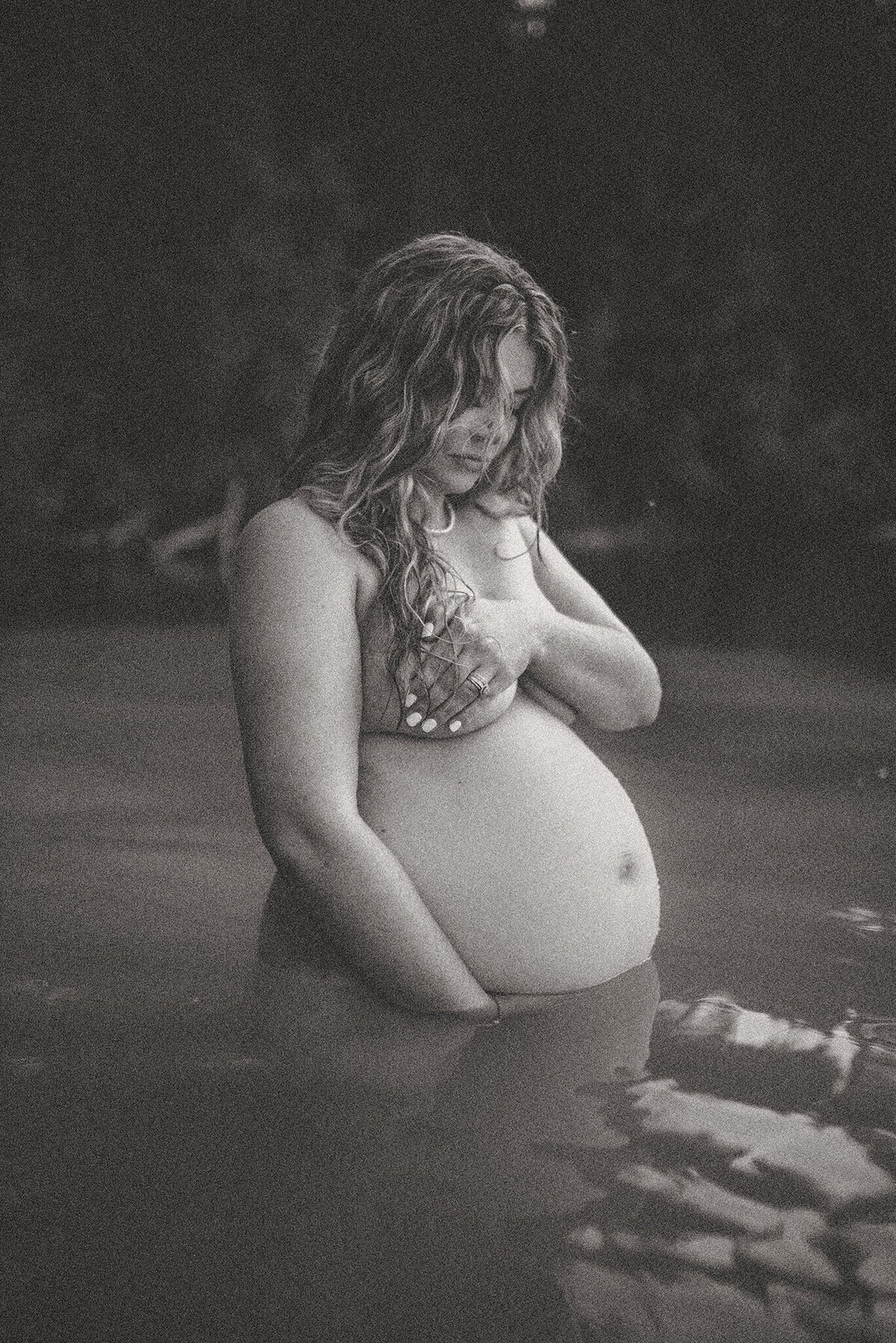 A naked pregnant woman is submerged to her waist in water while holding her belly. This black and white portrait captured by Brittany Danielle Photography.