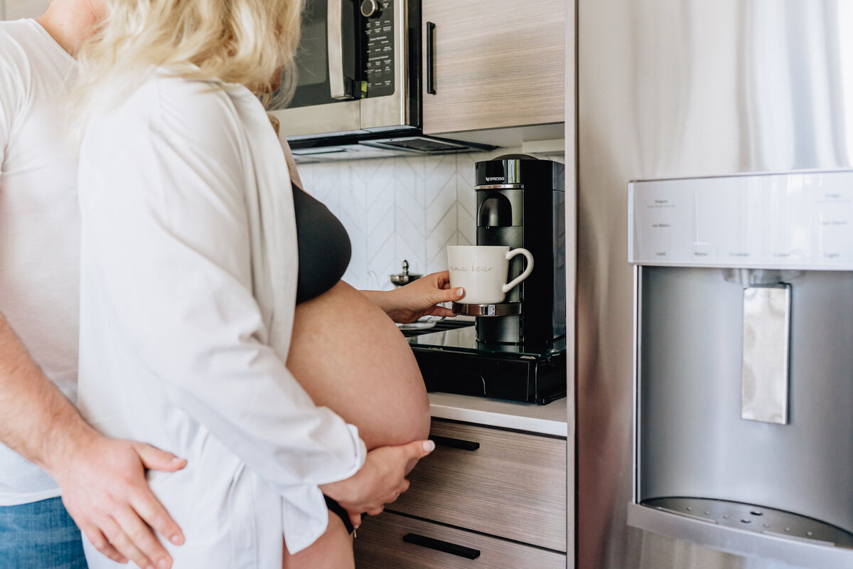A close-up of a pregnant woman holding a mug with "mama bear" written on it, as her partner embraces her from behind in the kitchen.