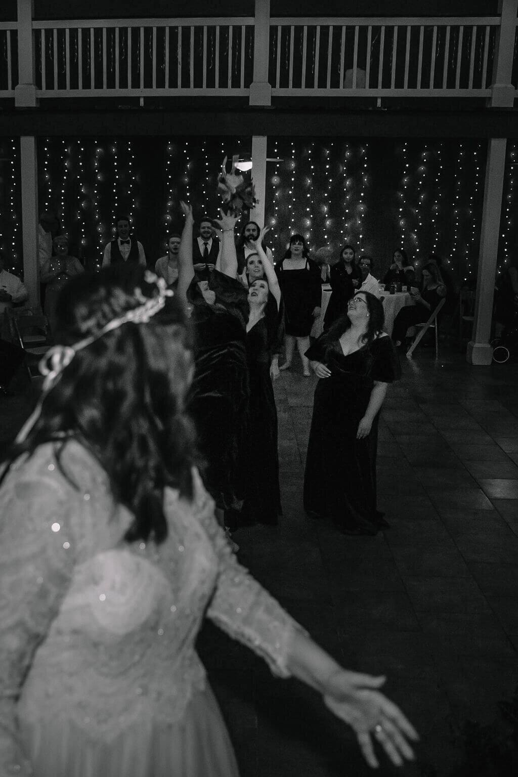 A bride in a sparkling dress tosses her bouquet to a group of women eagerly reaching to catch it, with fairy lights and seated guests visible in the background at a wedding reception.